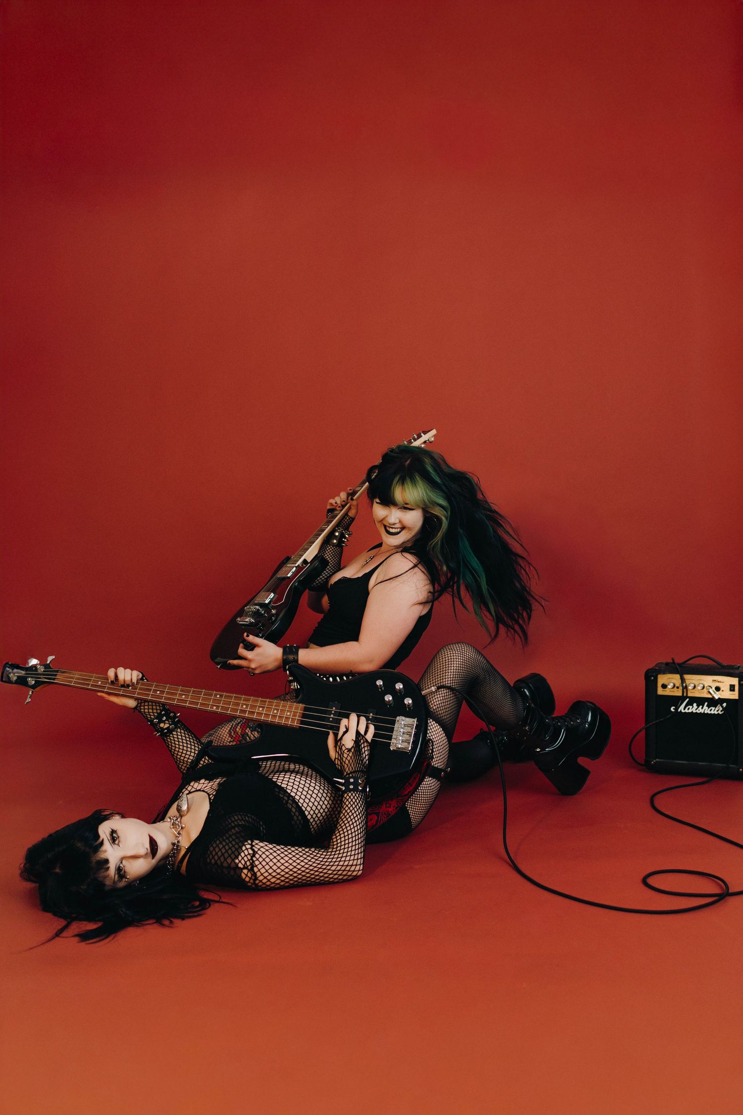 Two punk rock women pose with electric guitars on a red background, one lying down, beside a Marshall amp.