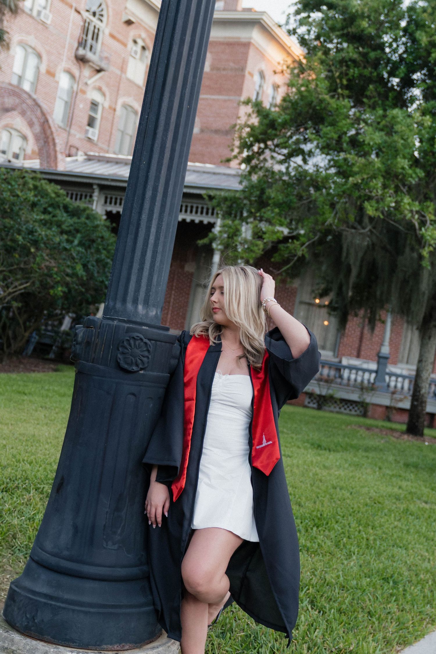 Blonde woman in black graduation gown with red stole leans against lamppost on college campus grounds.