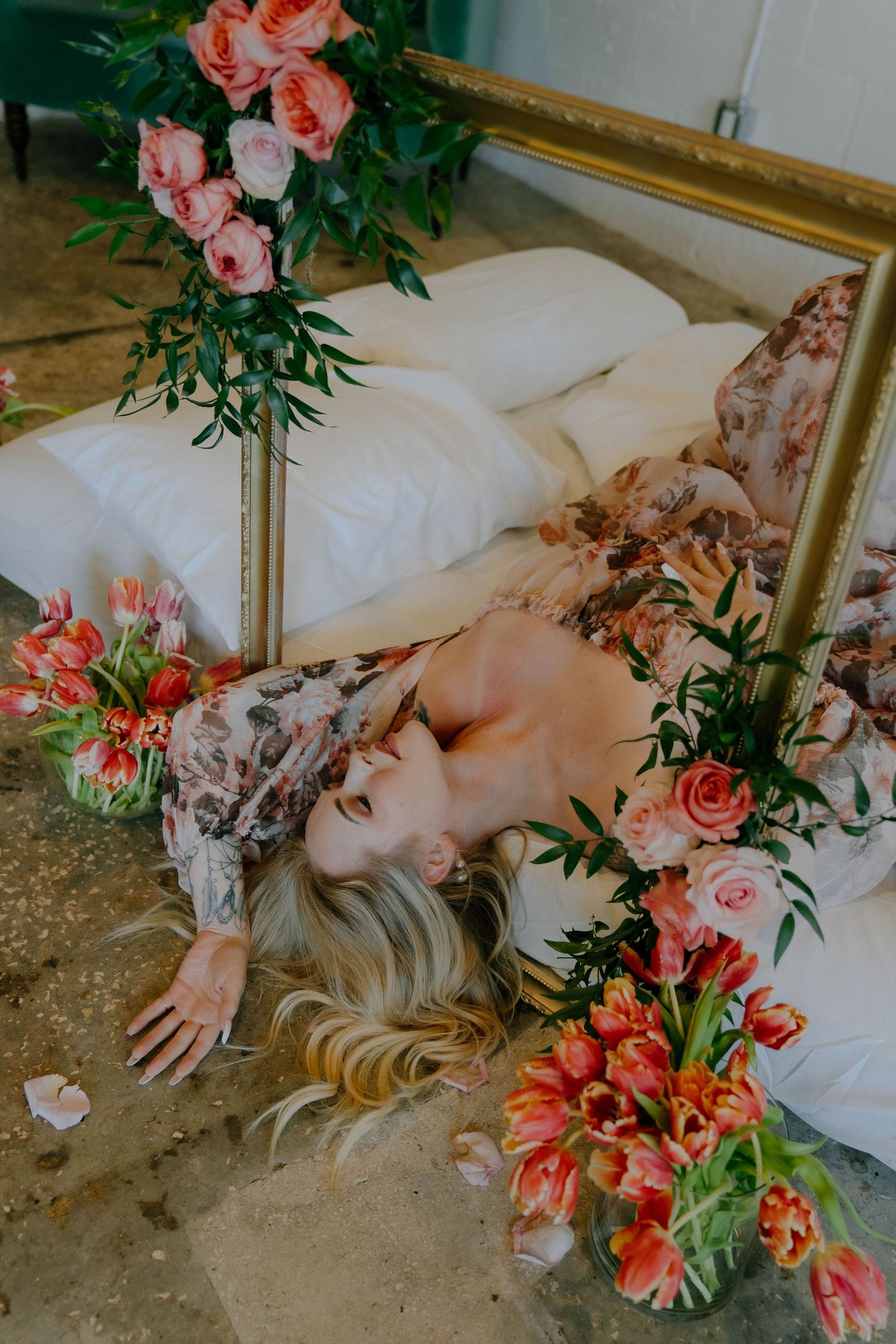 Romantic still life with coral roses and greenery arranged around a soft bed on a concrete floor.