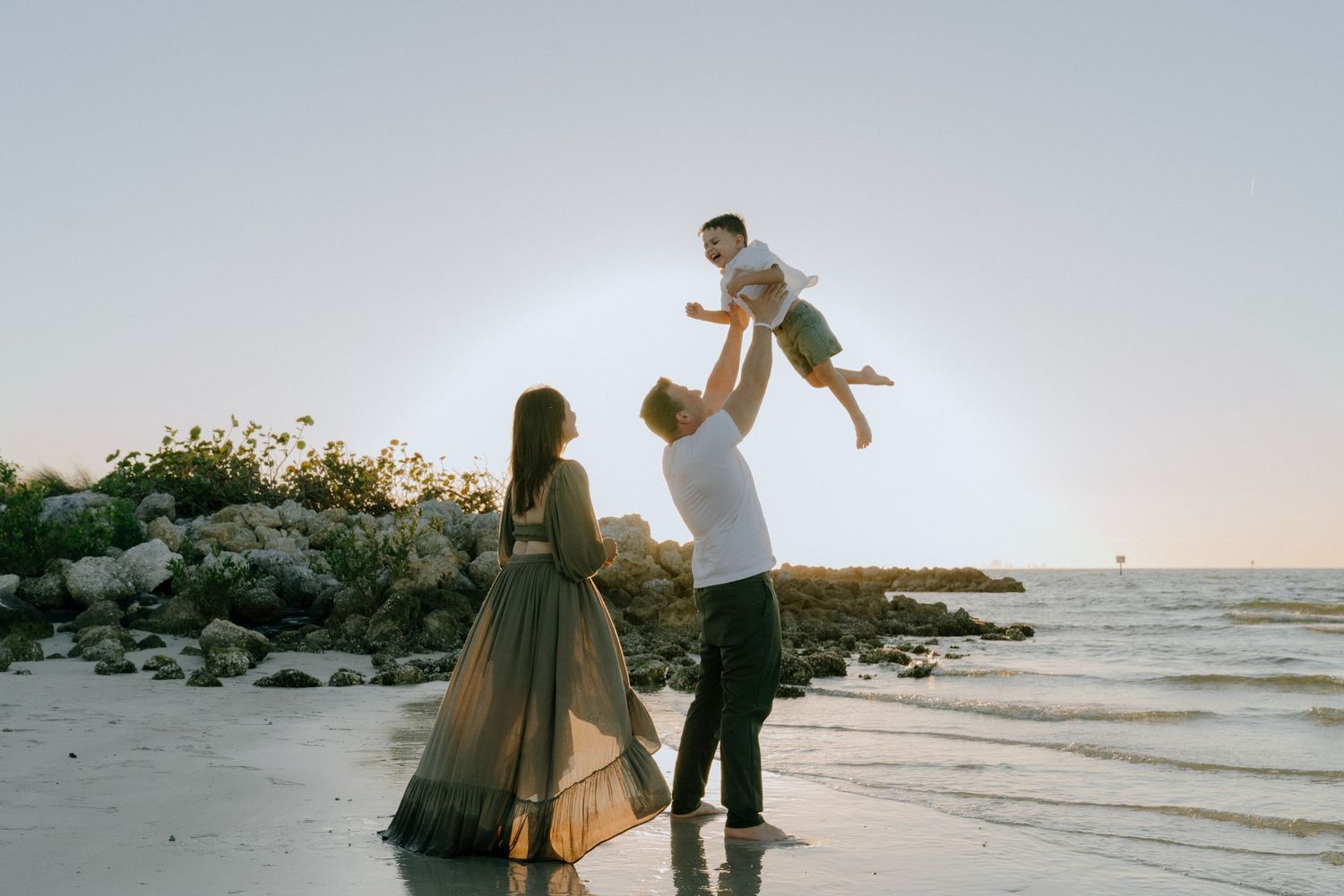 A couple and child spending time together at sunset on a beach with ocean waves in the background.