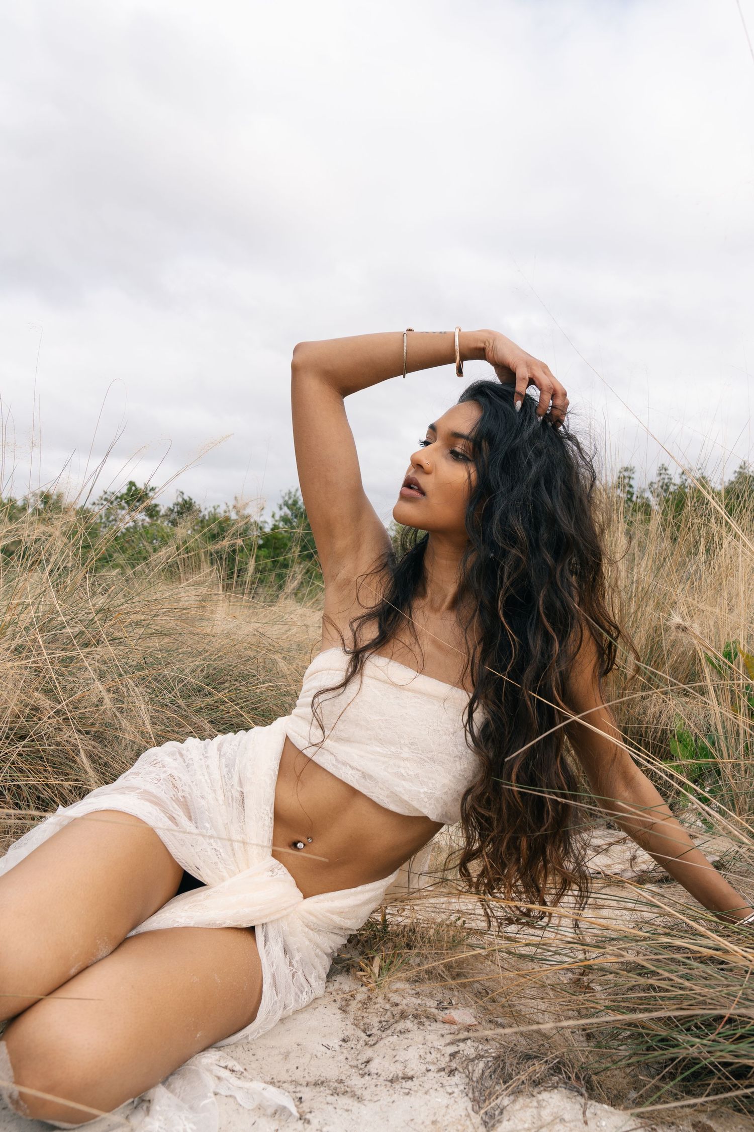 A woman in a white outfit poses dramatically amidst tall grass and sand dunes on a cloudy day.