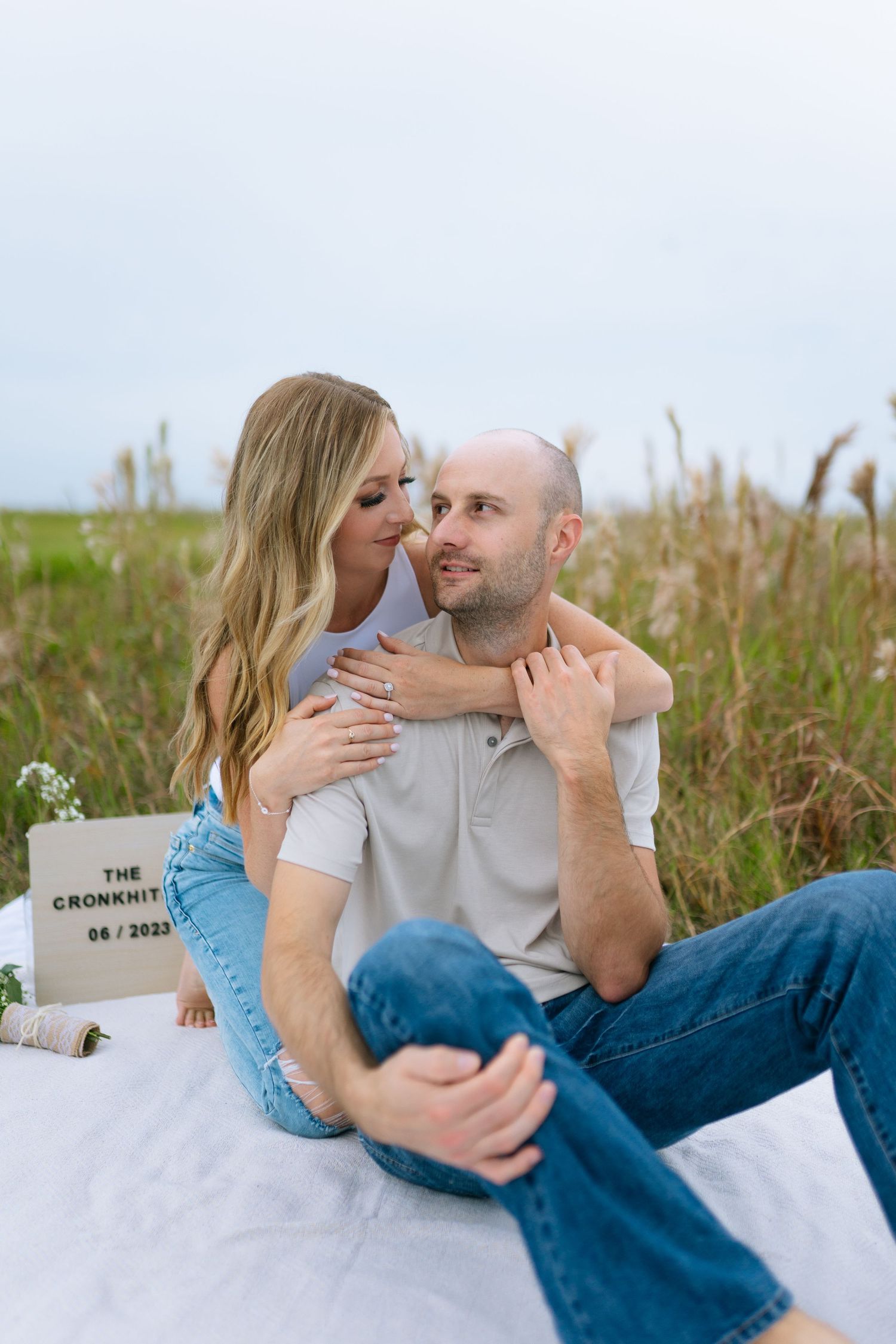 Intimate couple sharing tender moment while sitting on white blanket in tall grass field.