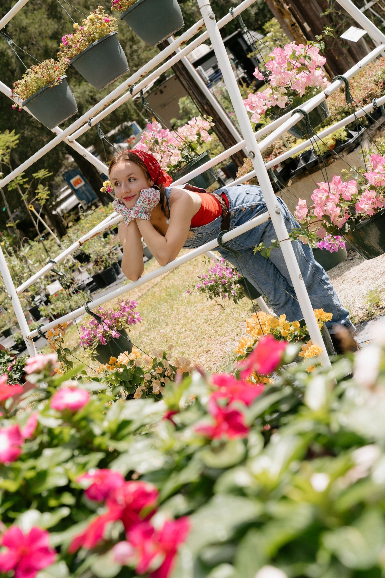 Young woman in red top and jeans leaning on white garden shelving surrounded by colorful blooming flowers outdoors.