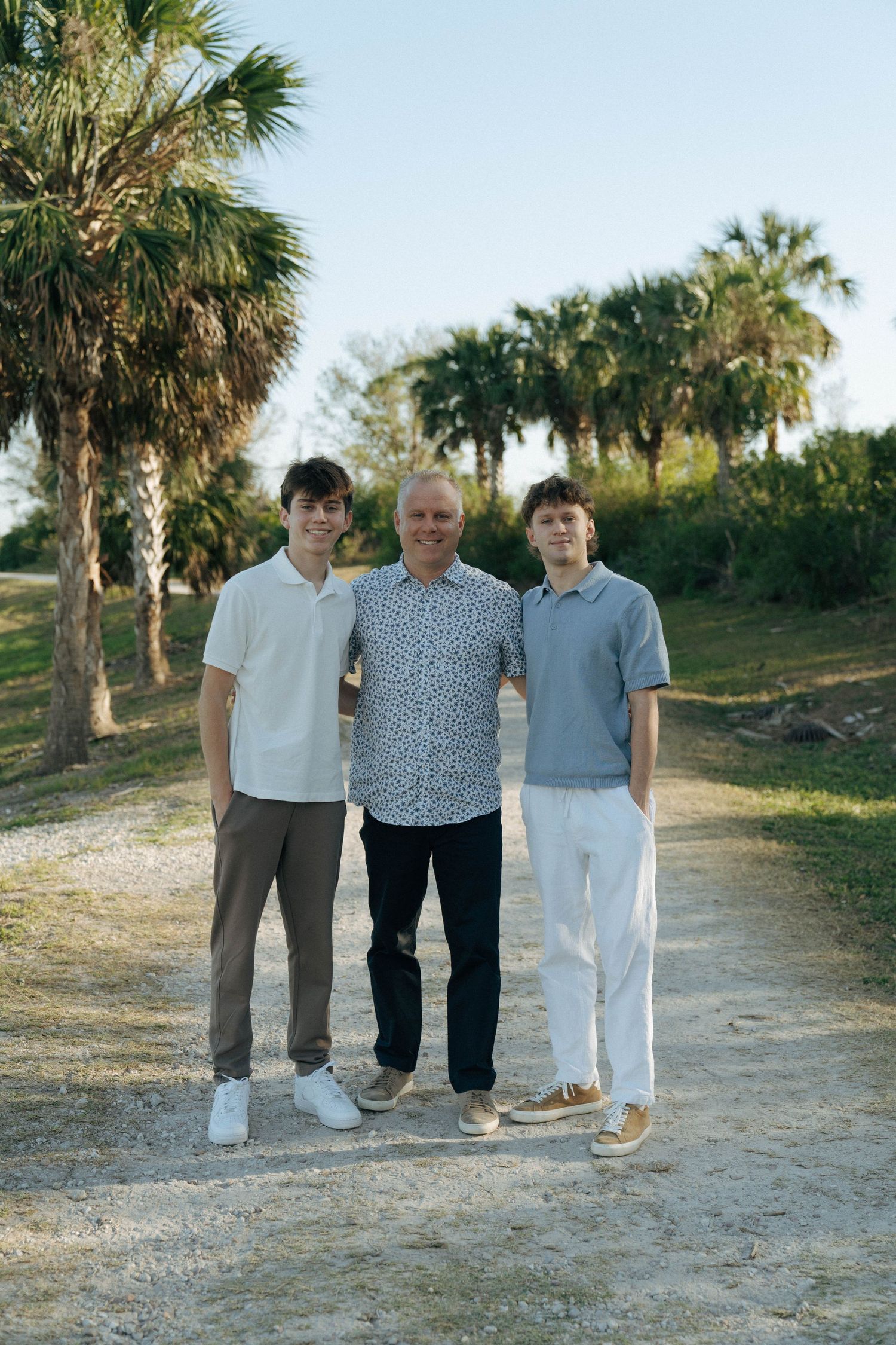 Three people stand together on a sandy path surrounded by palm trees in a sunny outdoor setting.
