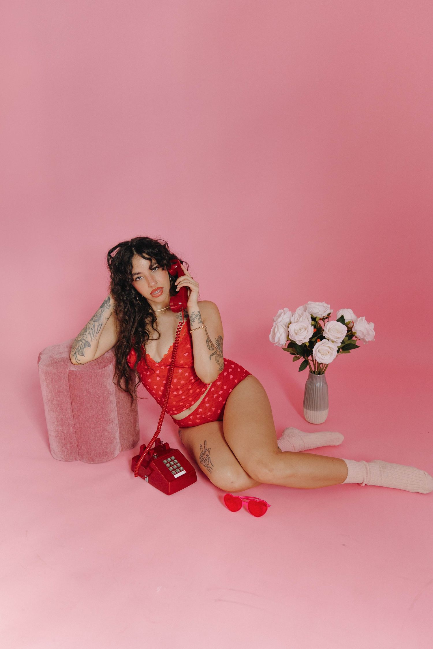 Woman in red polka dot lingerie sitting on pink floor with vintage phone, tattoos, white socks, and pink roses nearby.