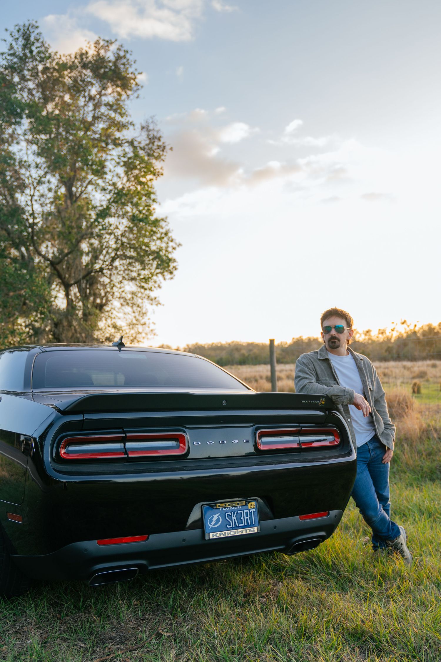 A black Dodge Challenger muscle car parked in a grassy field at sunset with trees in the background.