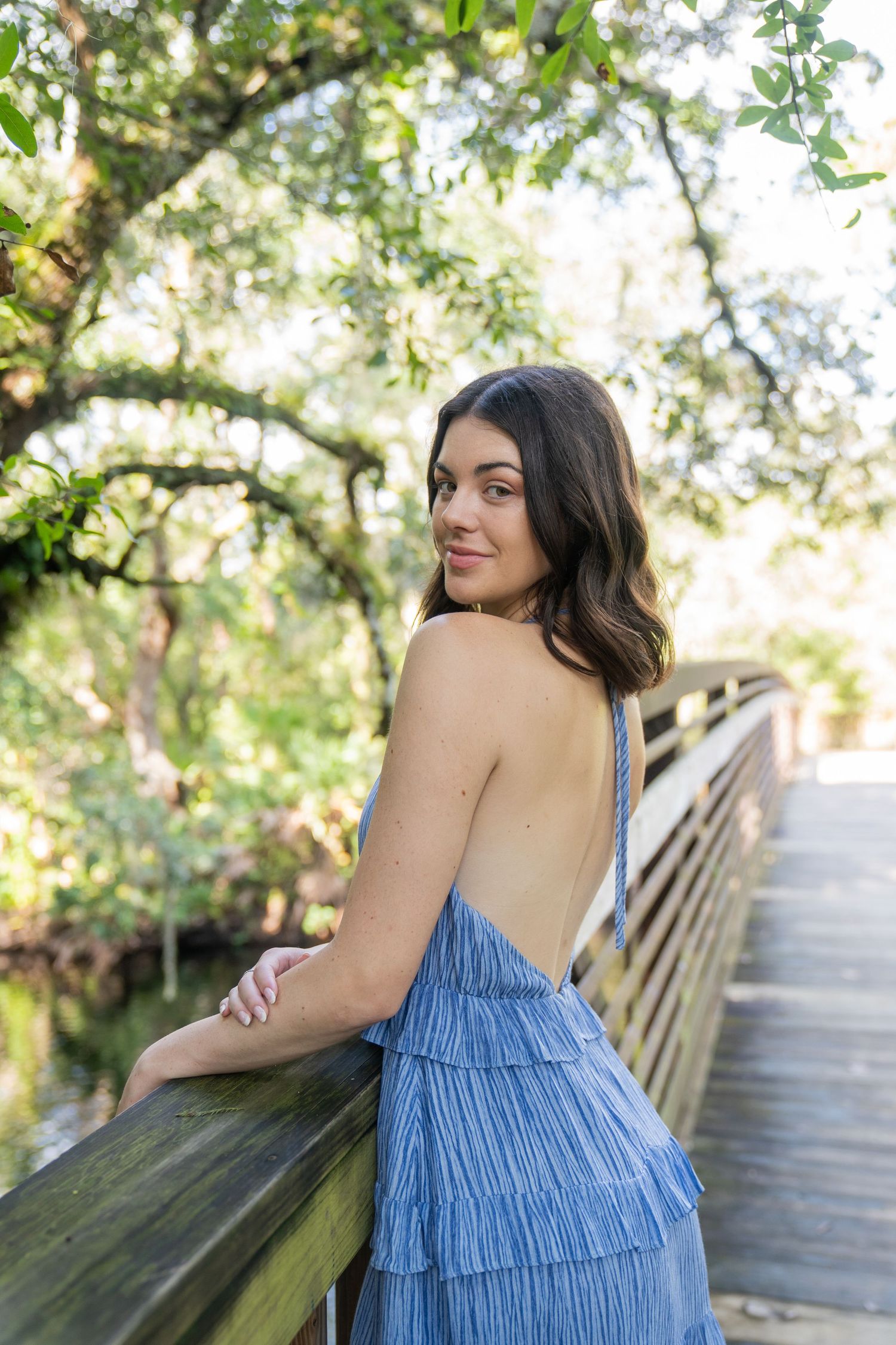 Woman in blue tiered dress poses on wooden bridge in natural setting.