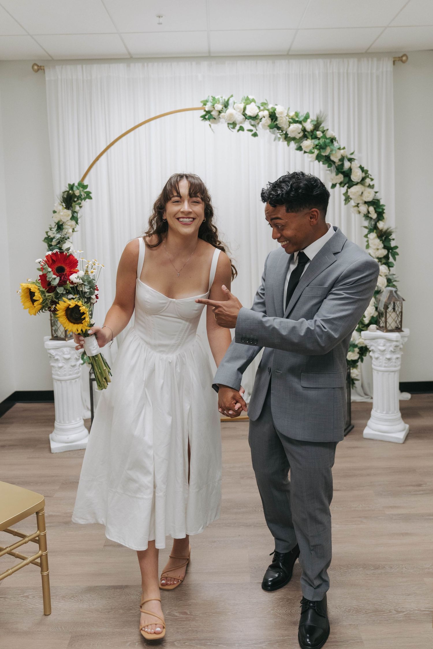 A couple dances together in a white wedding venue decorated with floral arches and sunflowers.