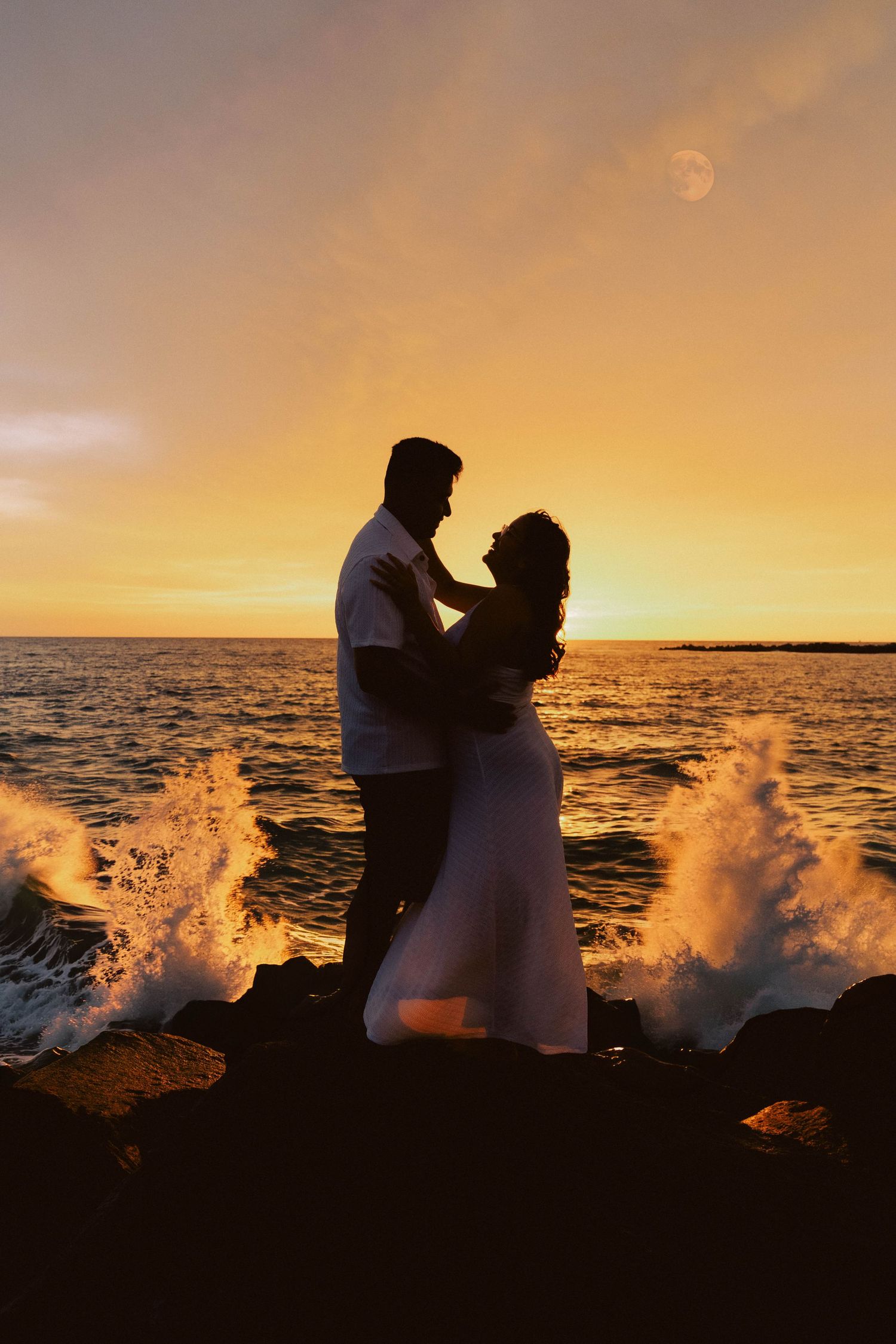 Silhouette of a romantic couple embracing on rocky shore at golden sunset with ocean waves crashing around them.