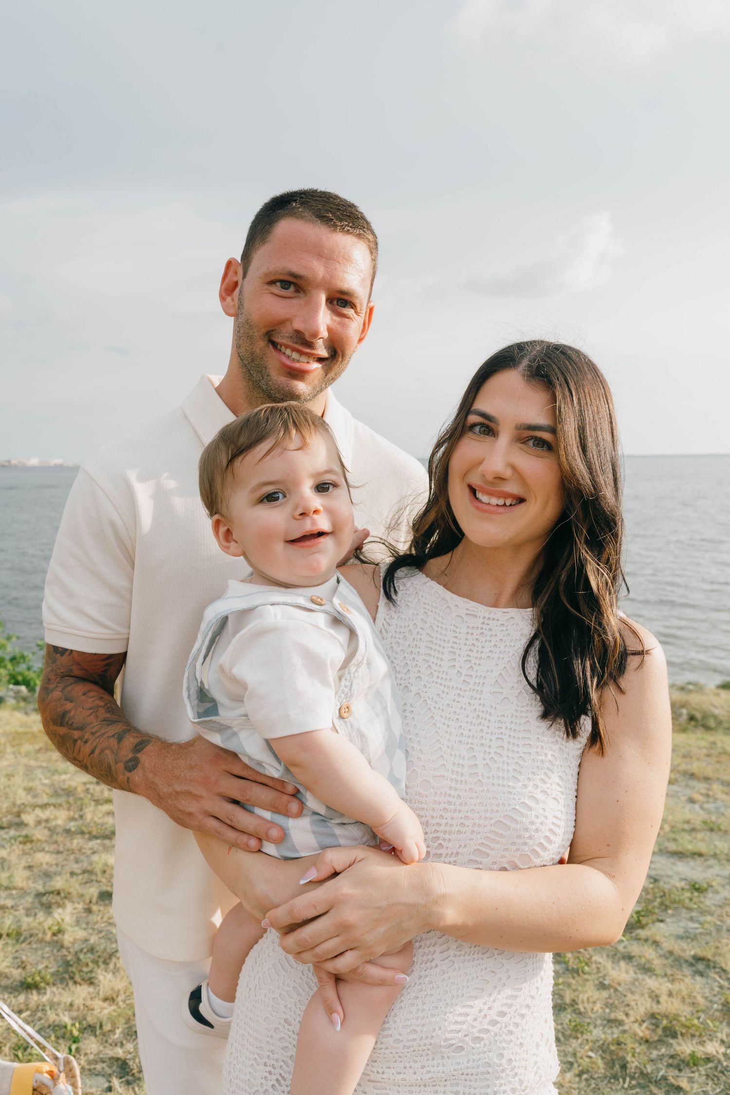 A smiling family of three poses outdoors near the water, with parents in white holding their happy baby boy.