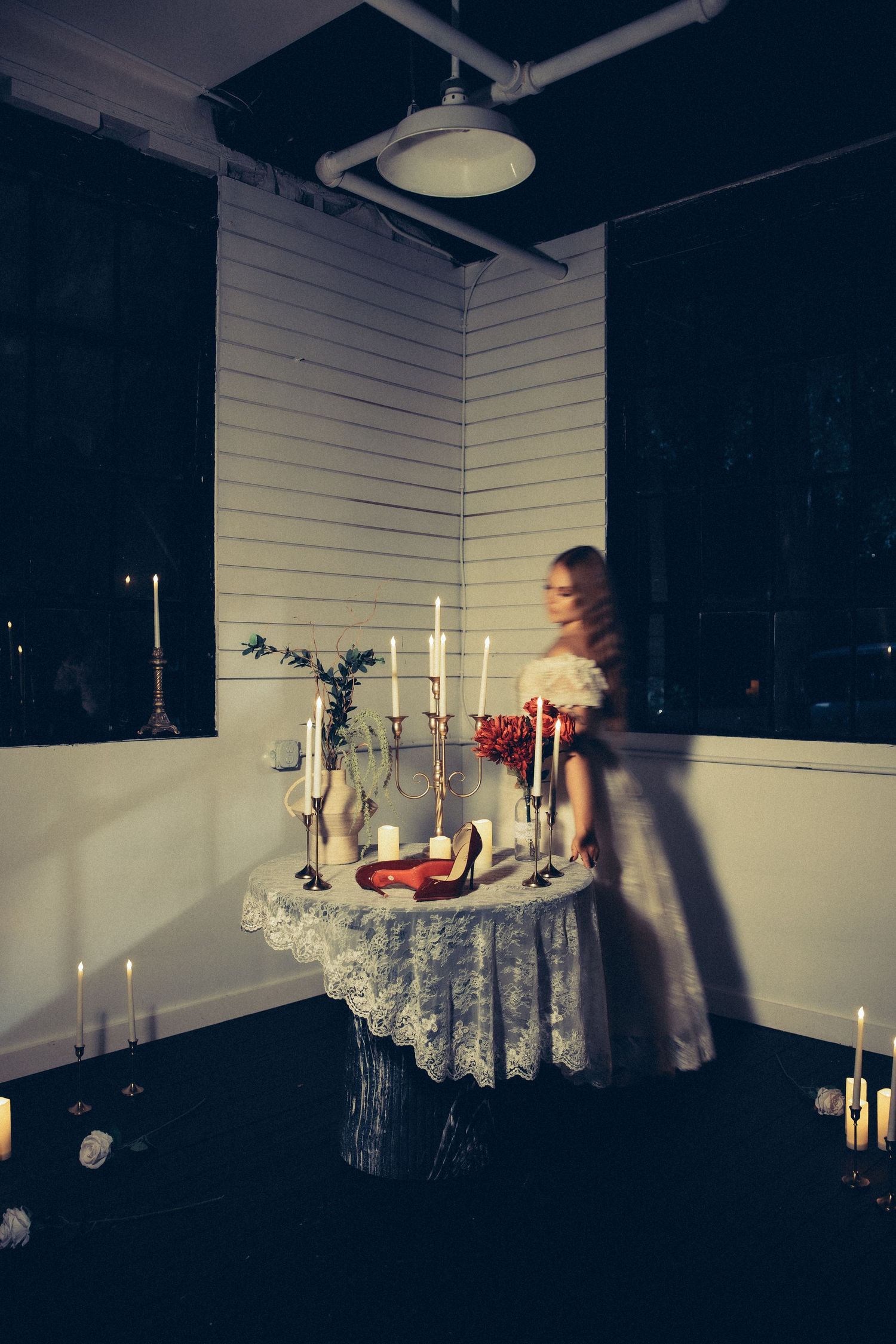 A blonde woman in white dress stands by candlelit lace-covered table with red heels and flowers in dark room.