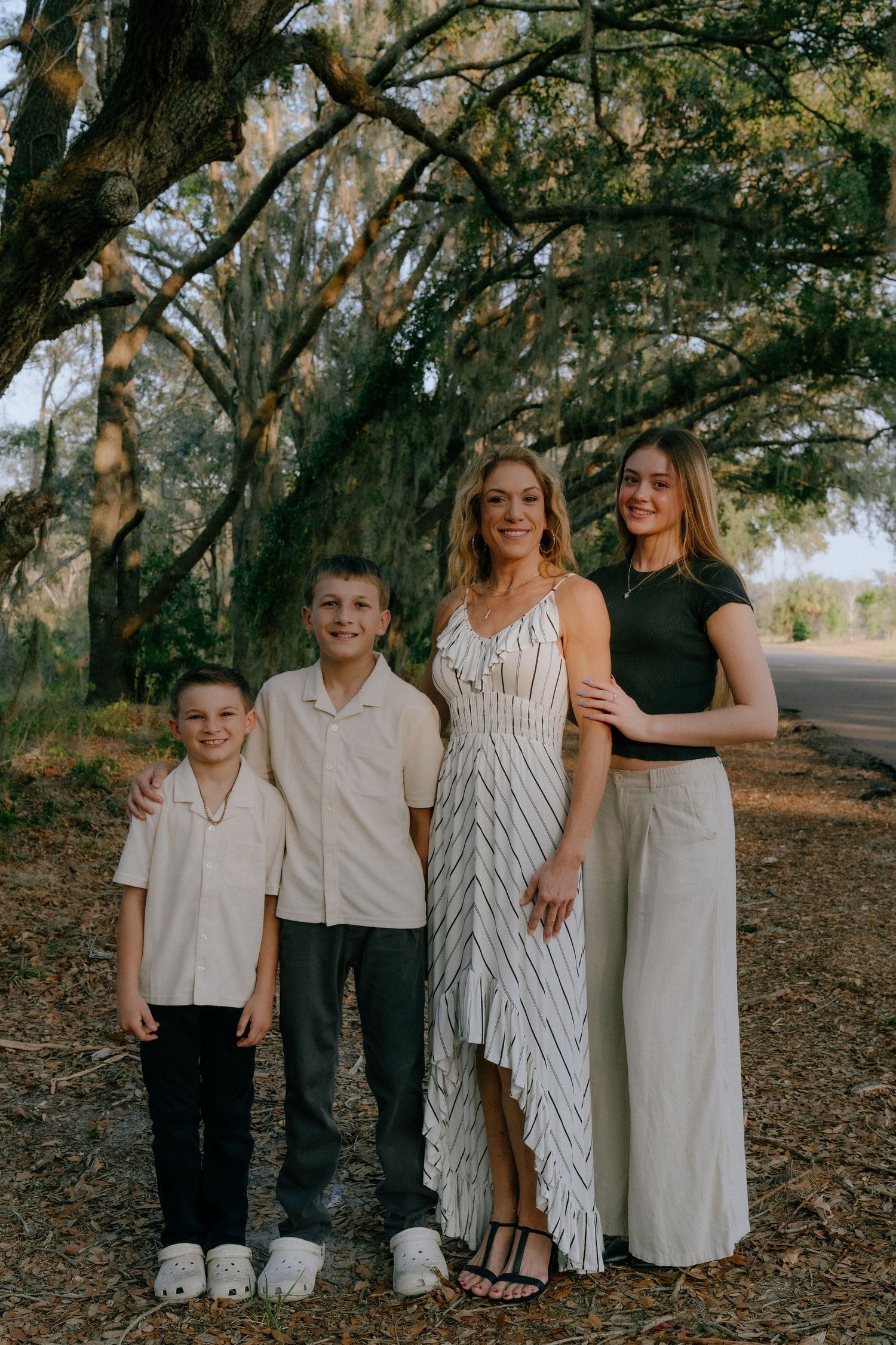 A family poses for an outdoor portrait beneath towering oak trees along a tree-lined path in casual coordinated attire.