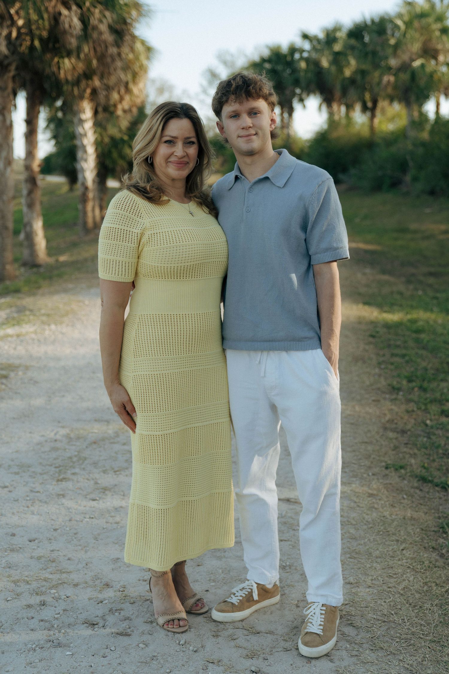 A couple stands together on a dirt path with palm trees, wearing casual summer outfits in yellow and blue tones.