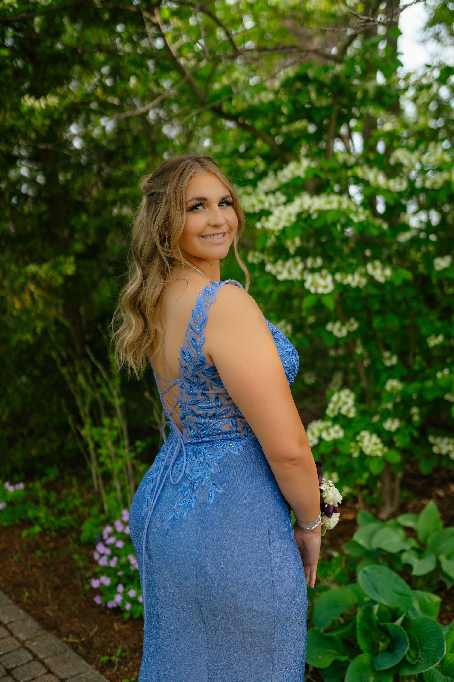 A person in a blue lace formal dress poses in a garden setting with flowering plants and greenery in the background.
