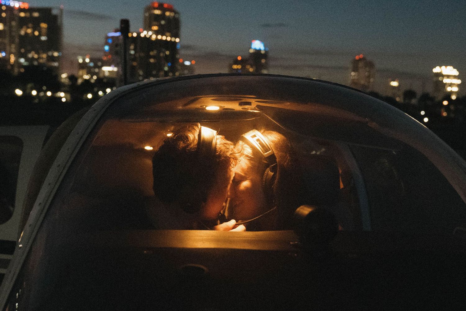 Couple sharing a romantic kiss inside a car at night with glowing city skyline in the background.