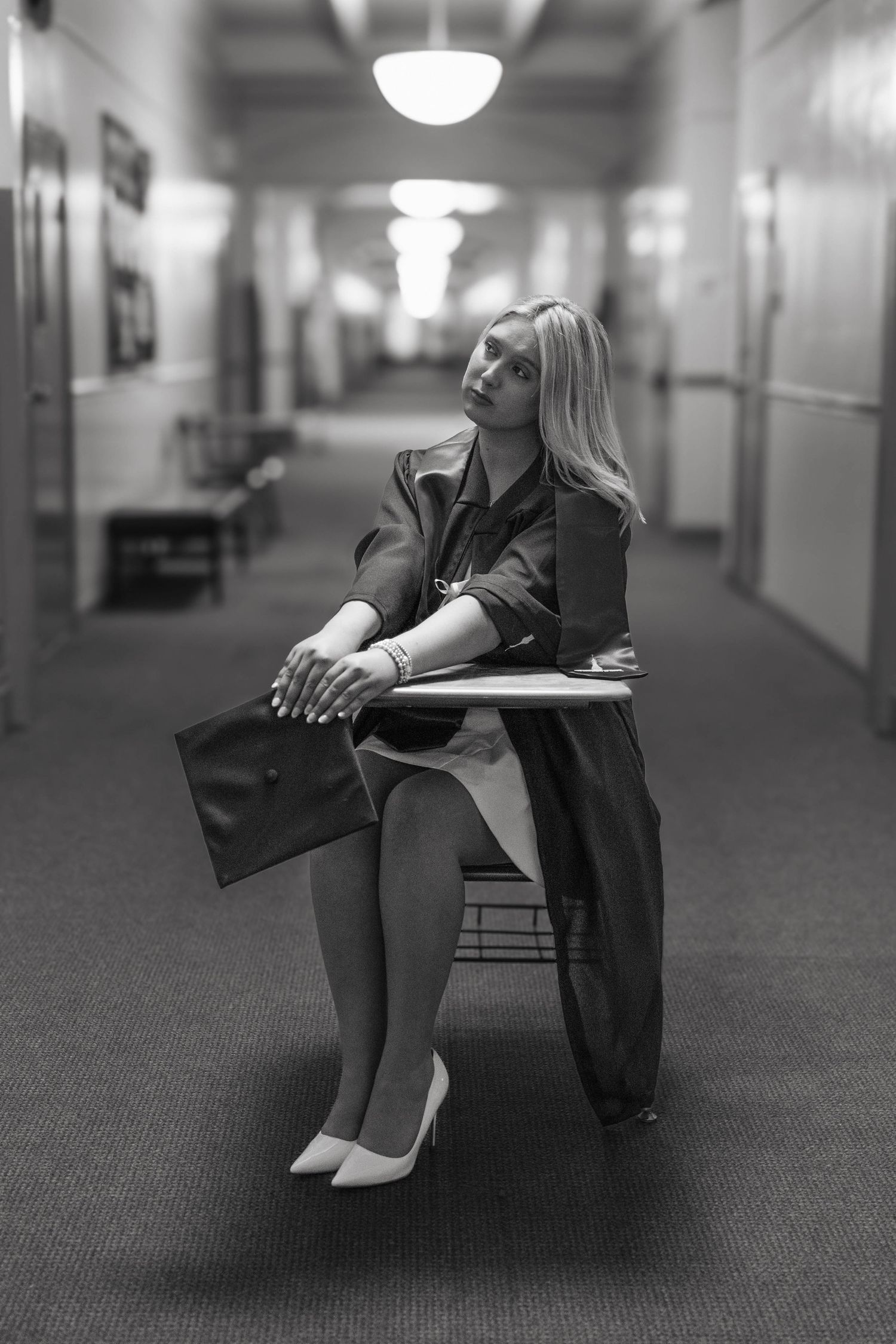 Black and white photo of a blonde woman in heels sitting in a school hallway chair, holding a graduation cap.