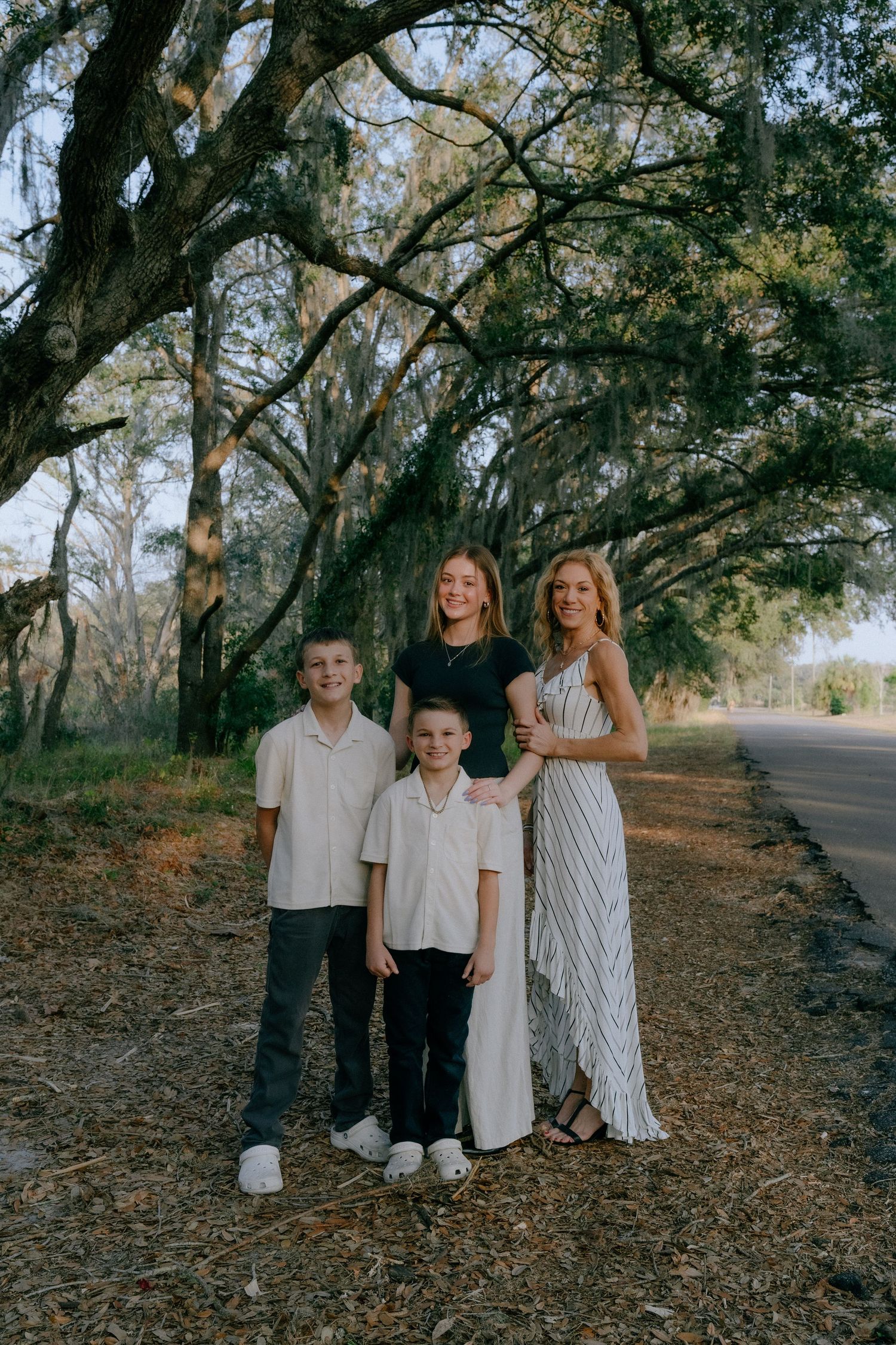 A family group stands together on a tree-lined path wearing coordinated white and neutral-colored outfits.