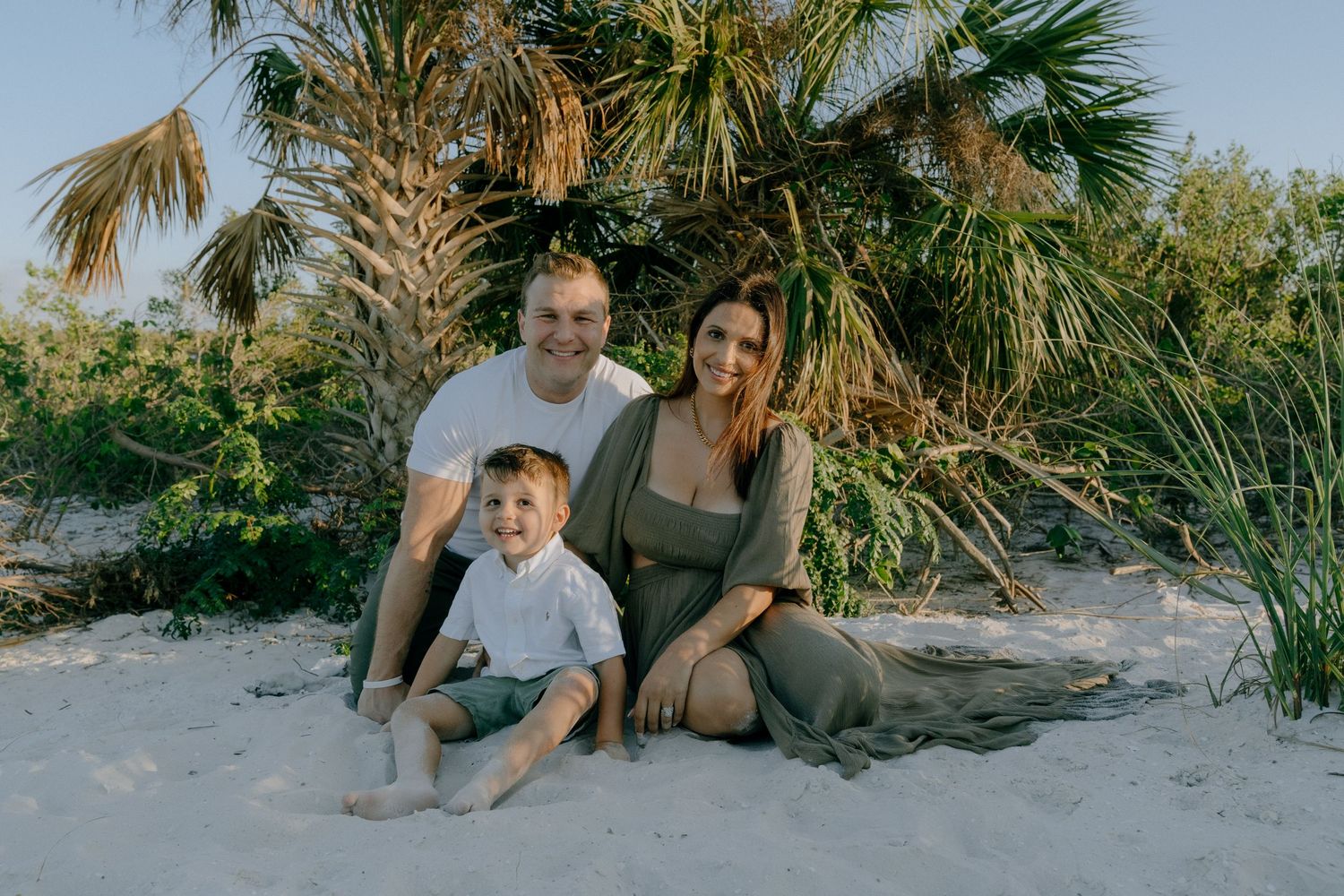 A family sits together on a sandy beach with palm trees in the background during golden hour.