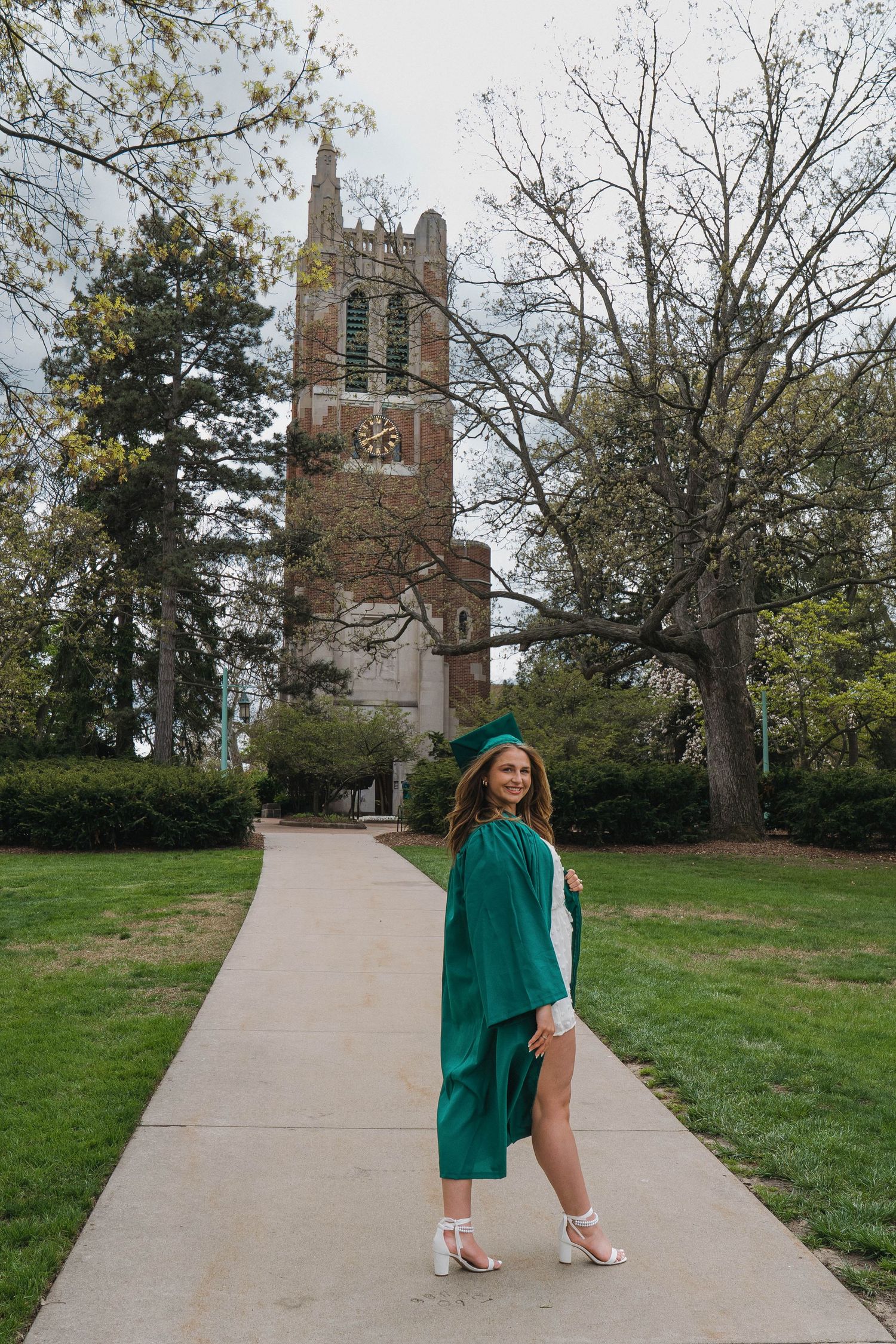 Smiling graduate in green cap and gown poses on campus walkway with historic brick bell tower in background.
