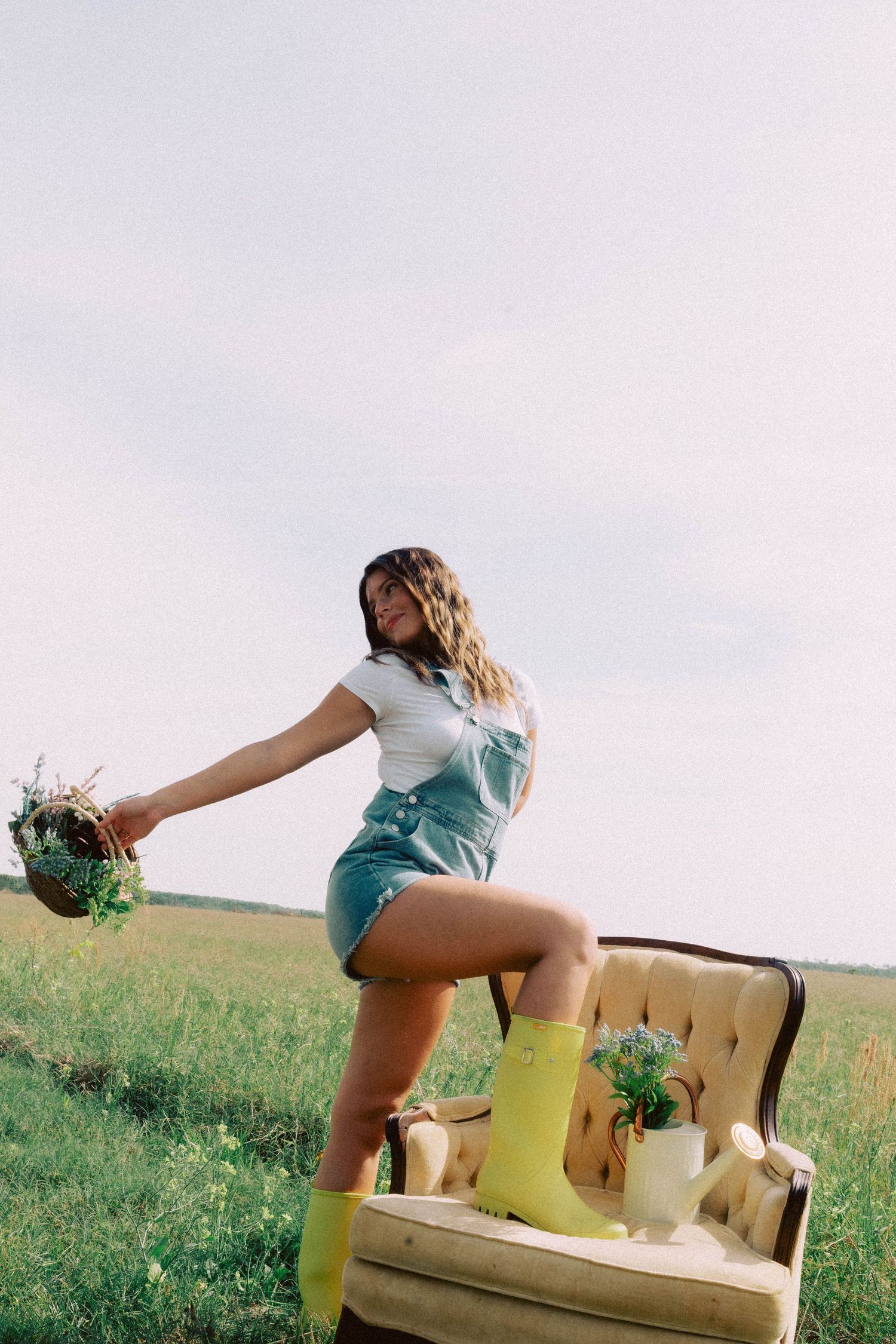 Young woman in denim overalls and yellow rain boots poses outdoors on vintage chair holding wildflowers in green field.