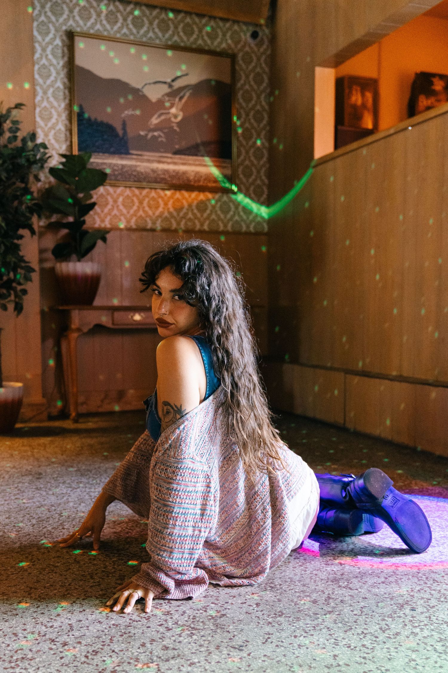 A person in a white lace dress and purple boots sits on a carpeted floor in a retro-styled room with ambient lighting.