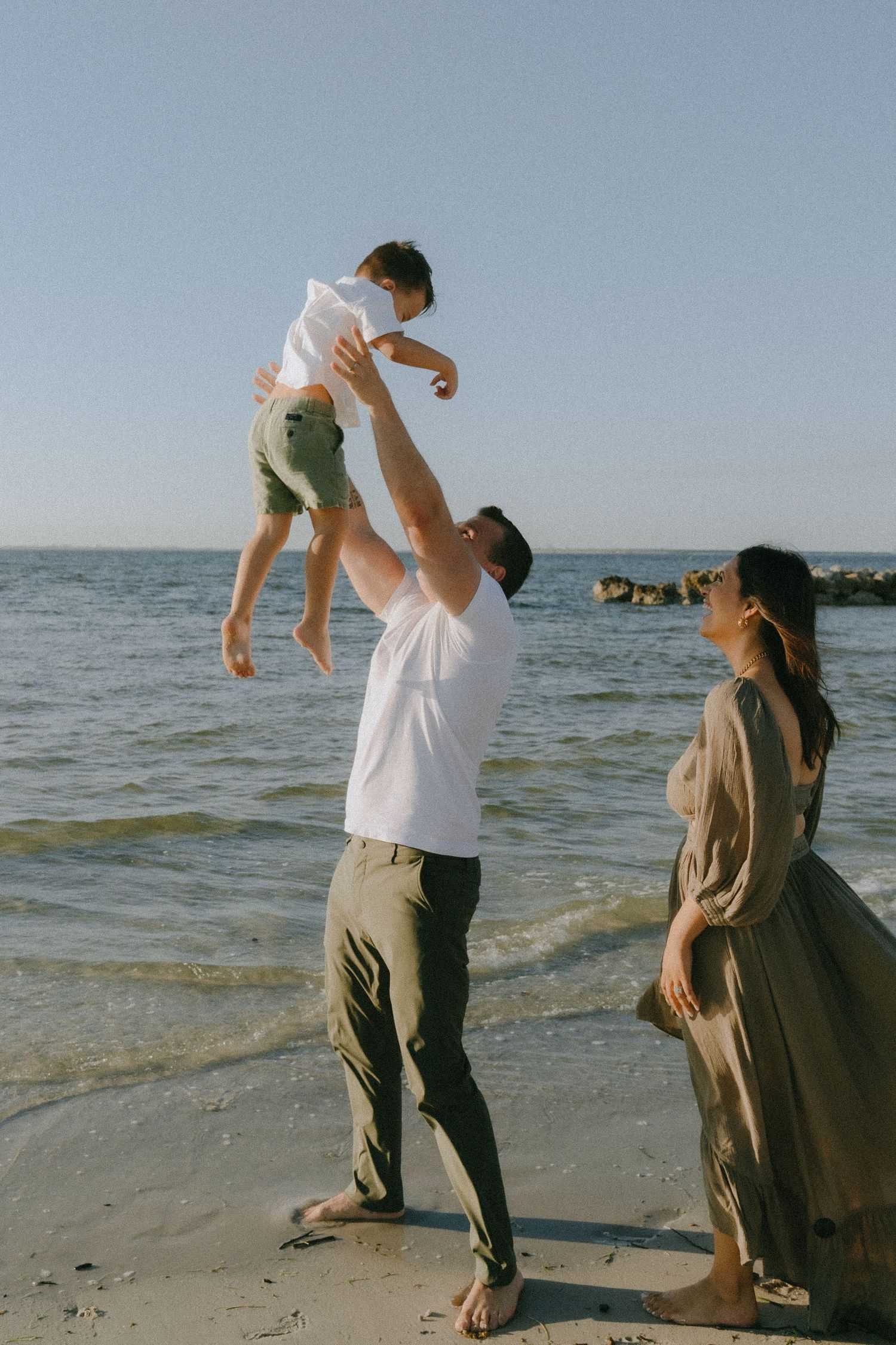 A family enjoys a playful moment together lifting a child up high on a sunny beach by the ocean.