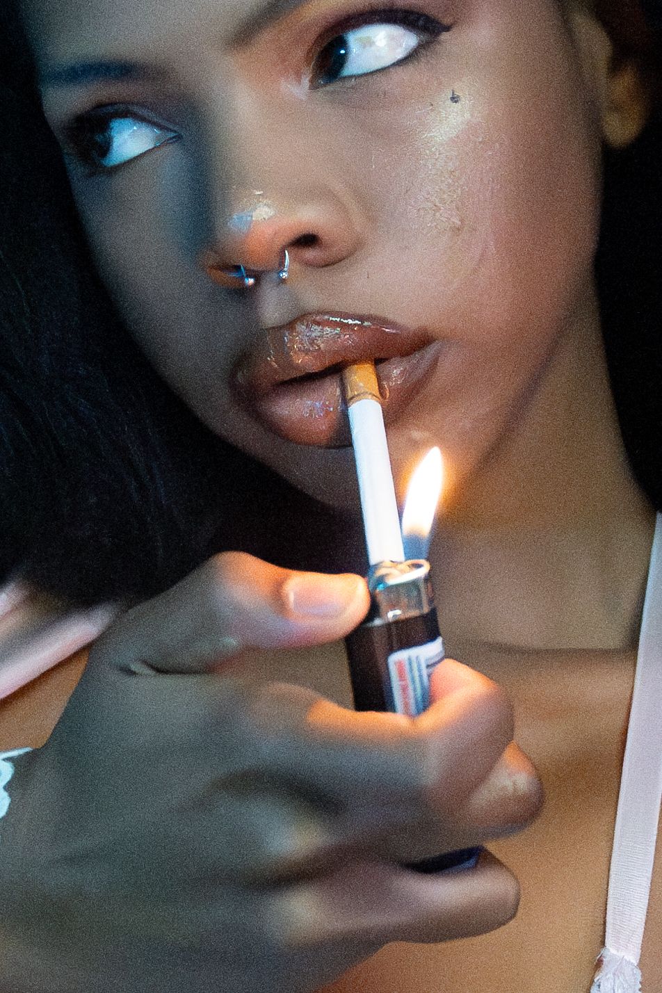 Artistic closeup photo of someone lighting a cigarette with dark lighting and pierced nose, showcasing glowing warm tones.
