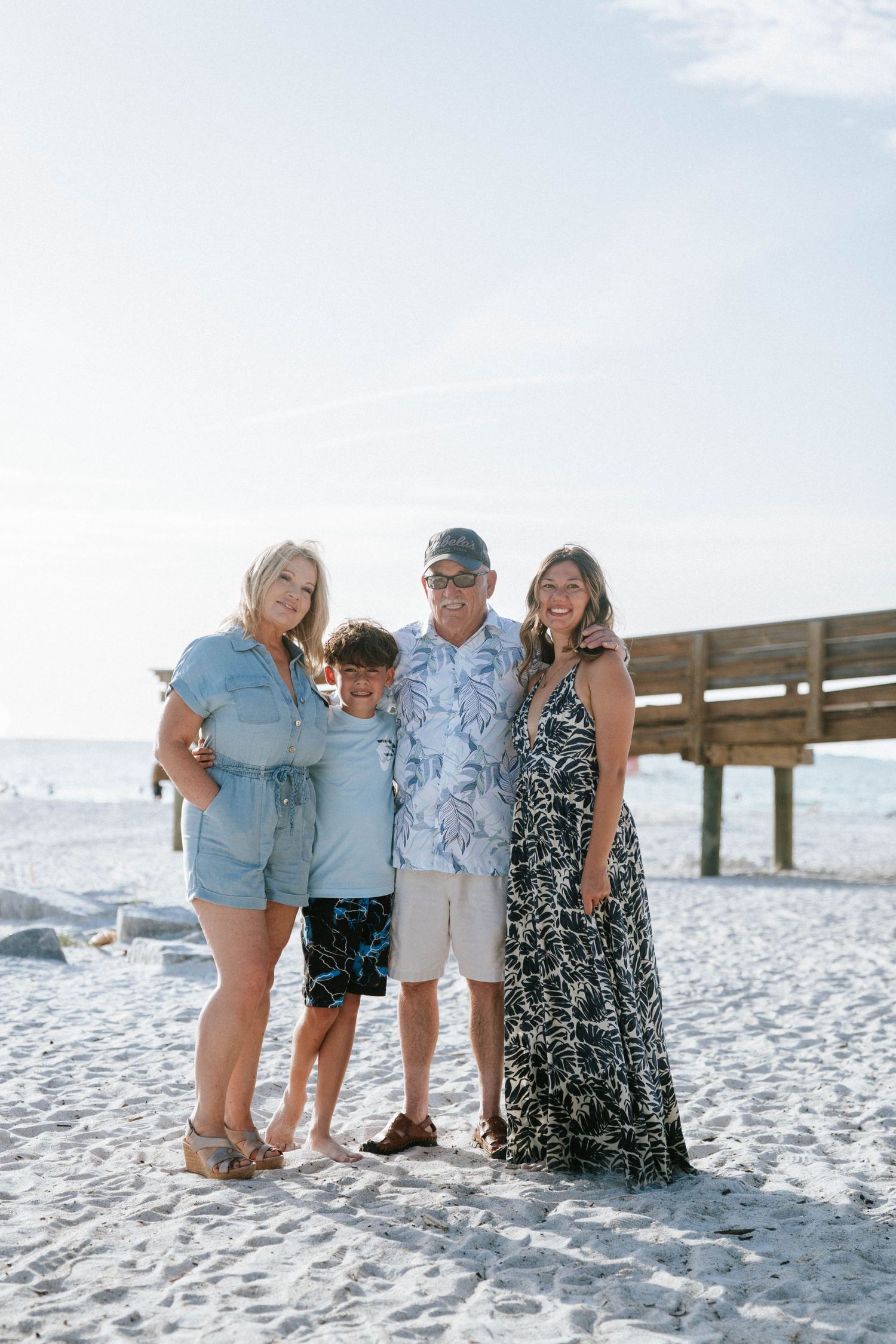 Casually dressed family standing together on a white sandy beach with a wooden pier in the background.