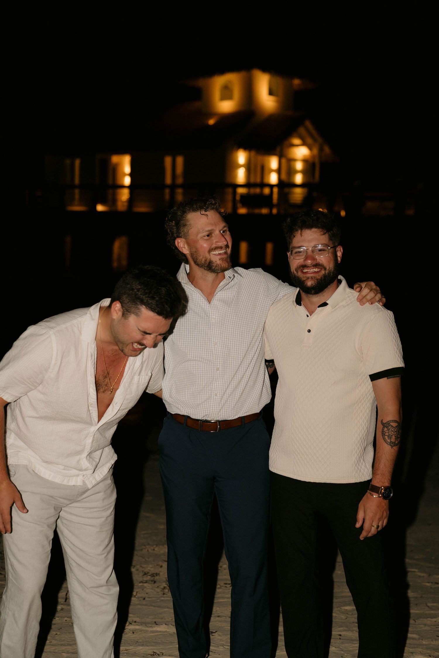 Three men smiling and posing together at night on a beach with illuminated building in background.