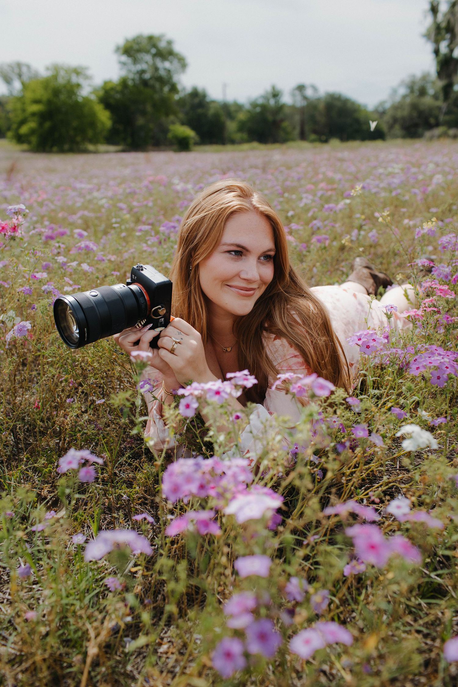 Young woman with red hair lying in a purple wildflower field holding a DSLR camera, smiling at the camera.