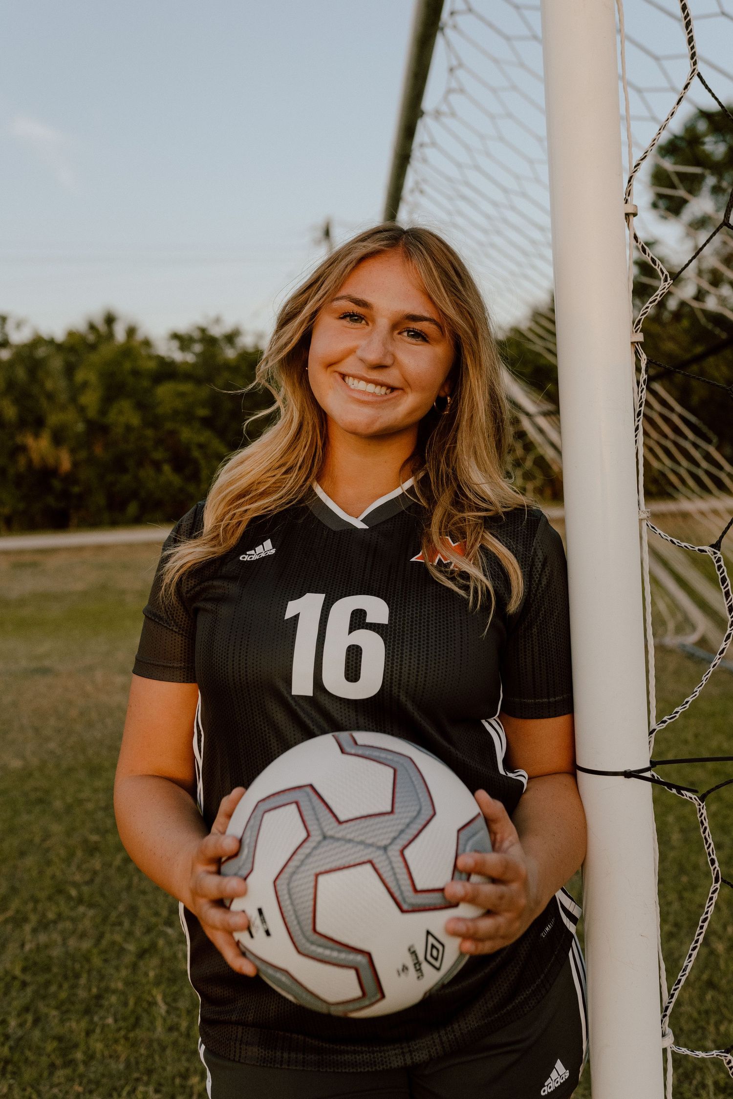 A soccer player wearing jersey number 16 holds a white ball while standing by the goal post at sunset.