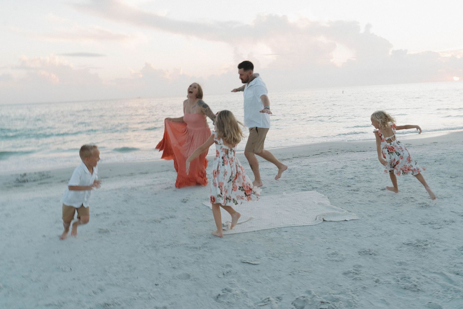 Family of five playing on a sandy beach at sunset, with children running and parents holding a flowing red scarf.