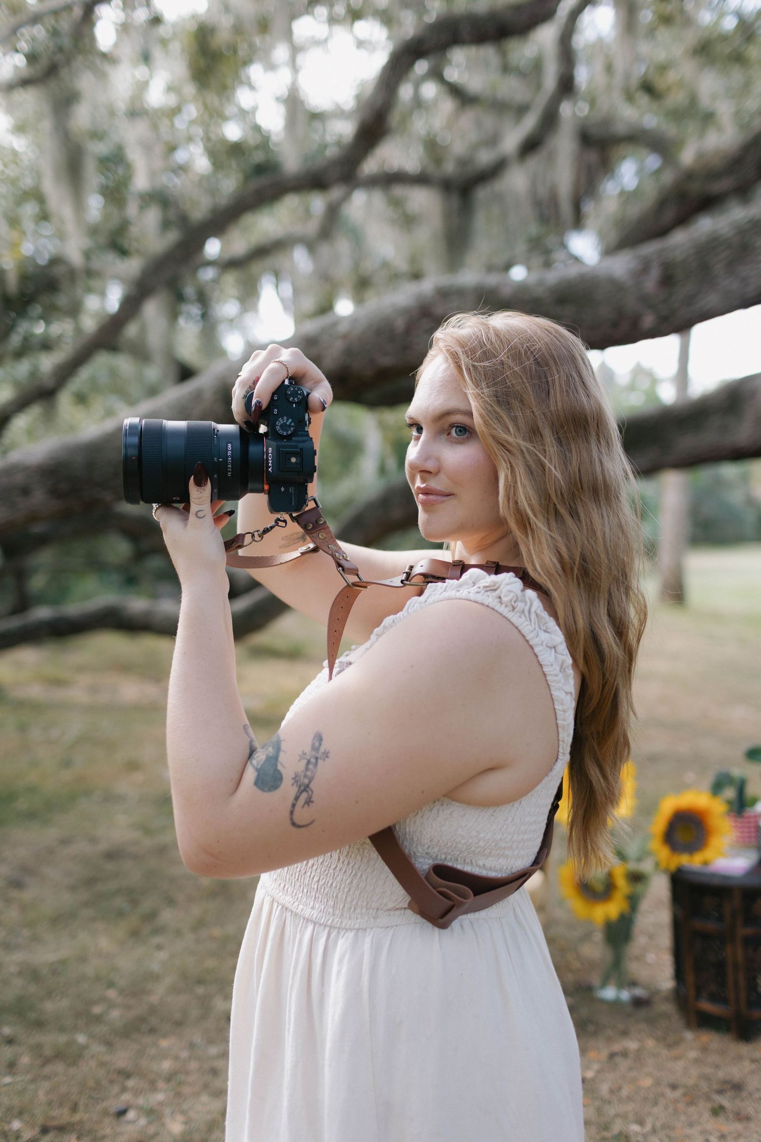 A woman in a white dress holds a DSLR camera outdoors near a large tree with sunflowers in the background.