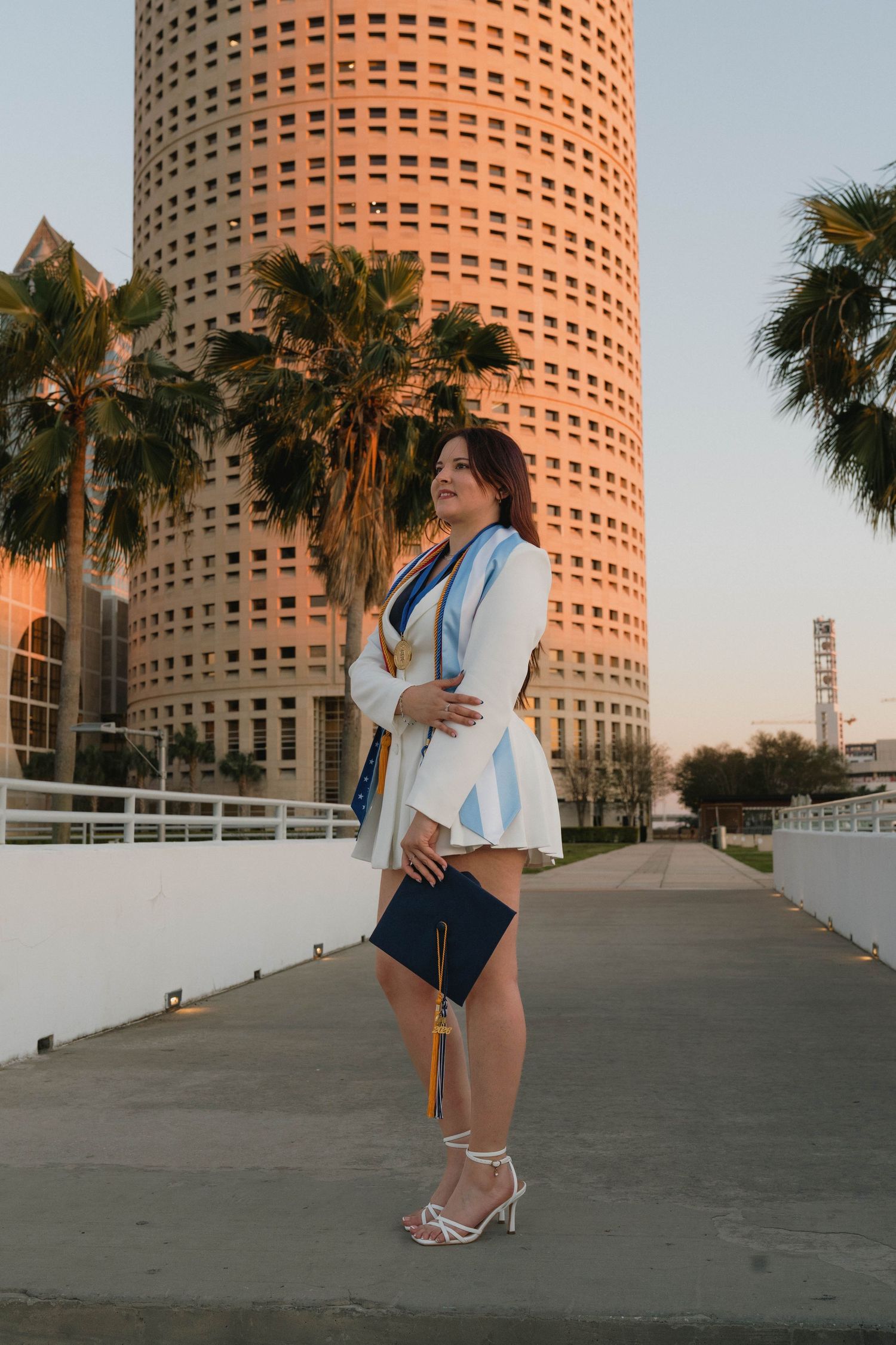 Graduate in white blazer holding cap near Tampa skyline with palm trees at sunset graduation portrait.