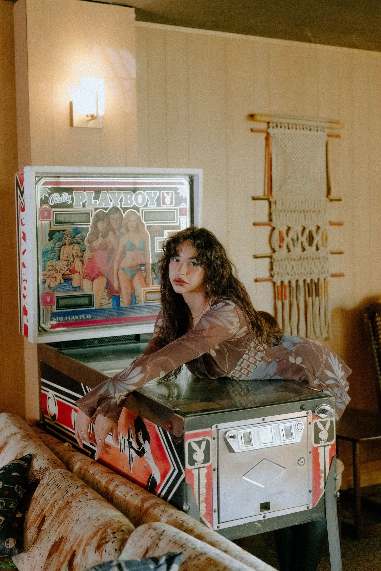 Woman leaning on a vintage Playboy pinball machine in a retro-styled room with macrame wall decor.