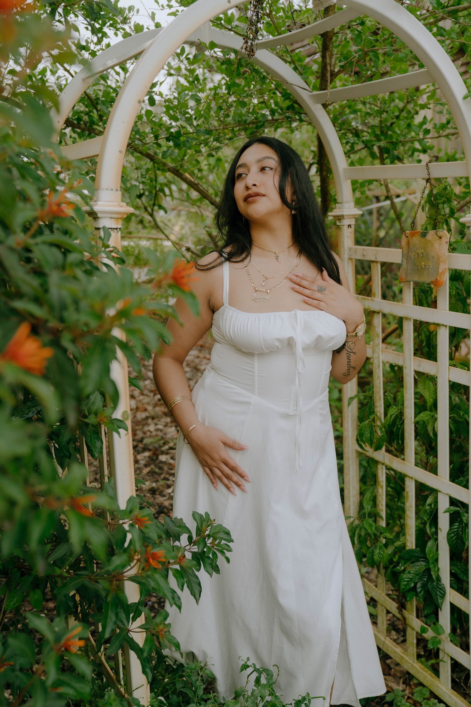 A person in a white sundress poses beneath a white garden arch surrounded by lush greenery and orange flowers.