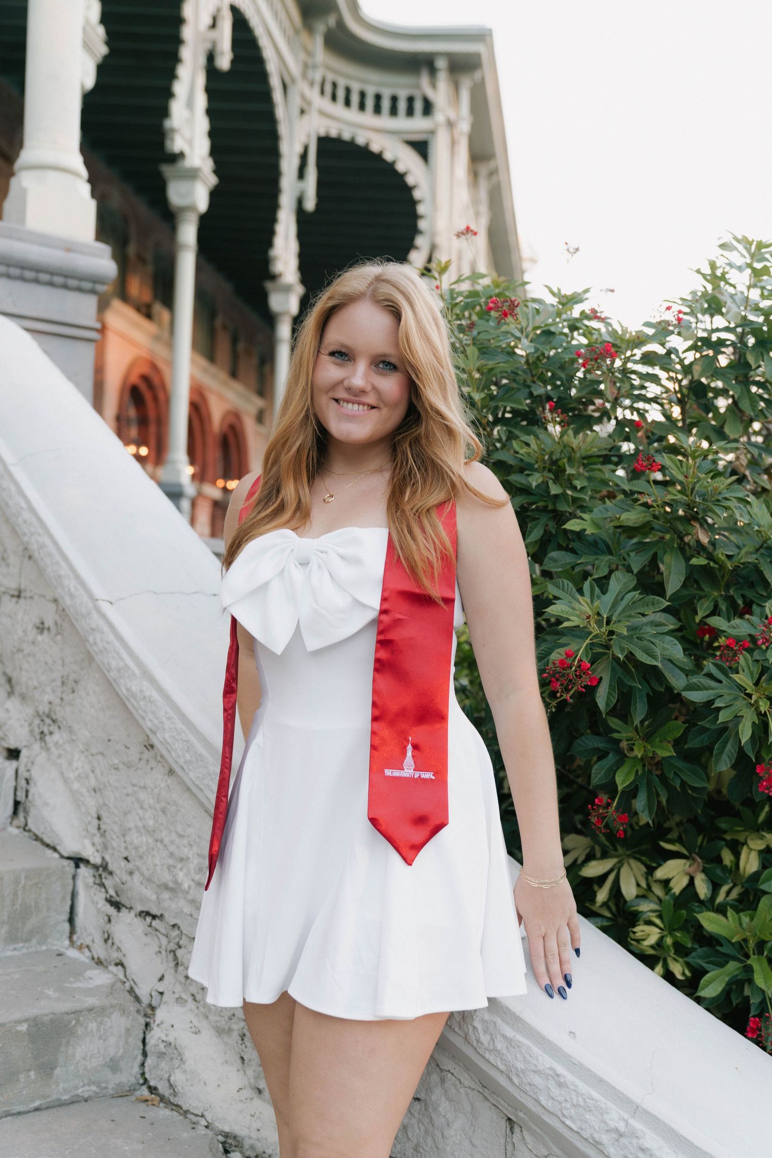 Young woman in white dress with red graduation stole smiling on stone steps near historic building.