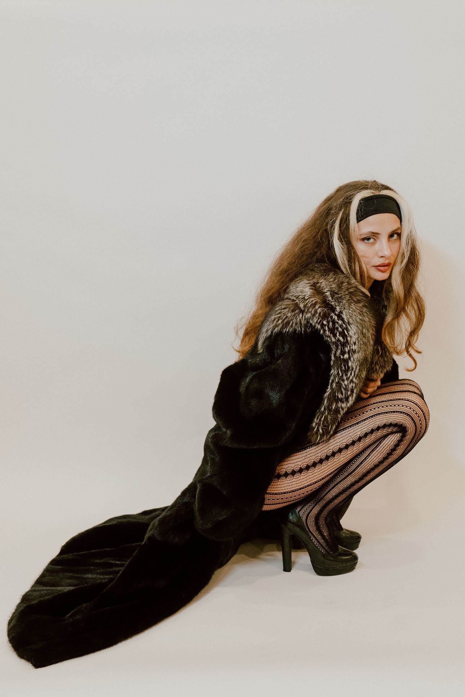 Fashion model poses in vintage fur coat and patterned tights against white backdrop in retro-inspired photoshoot.