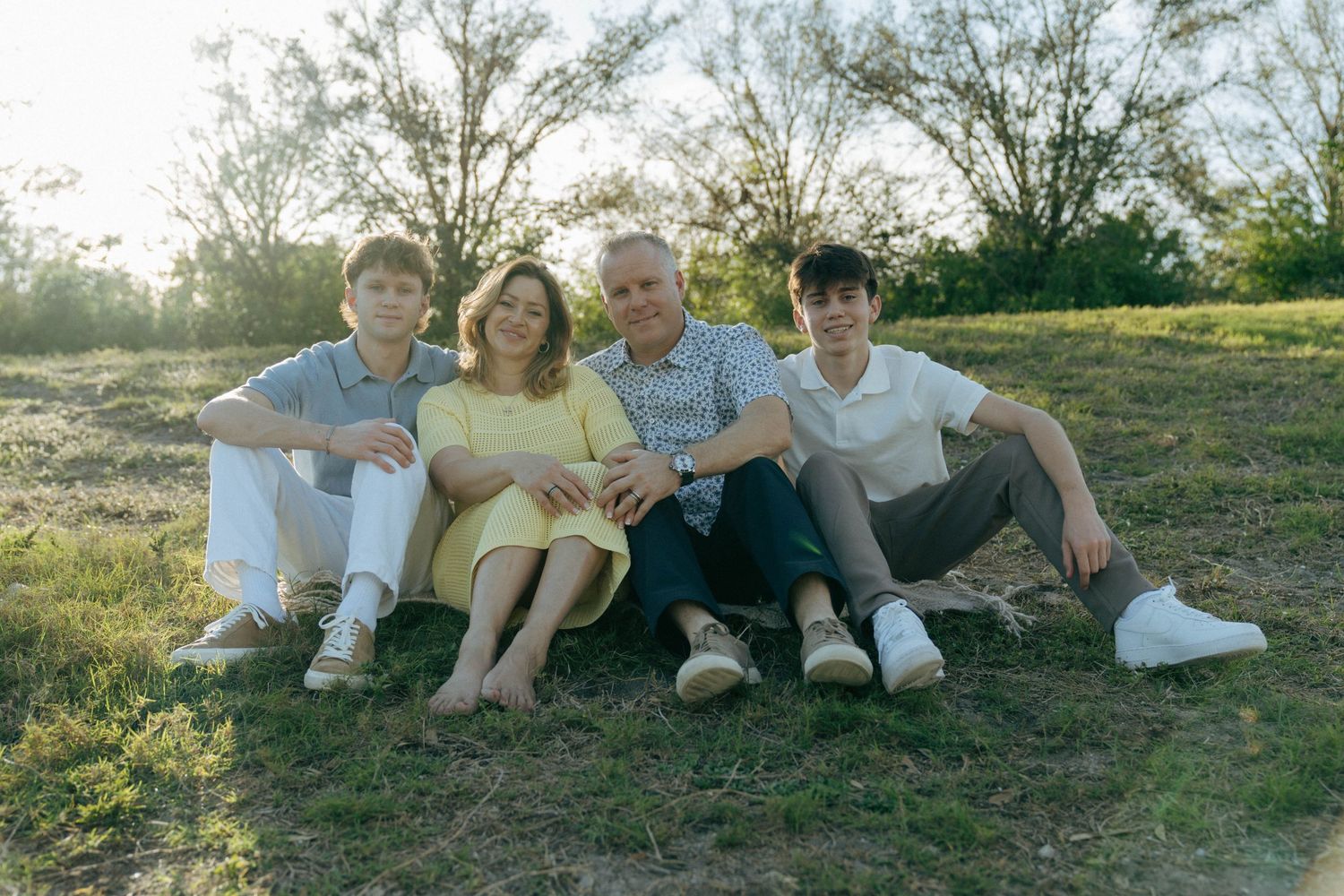 A family sits together on grassy ground during golden hour with trees in the background.