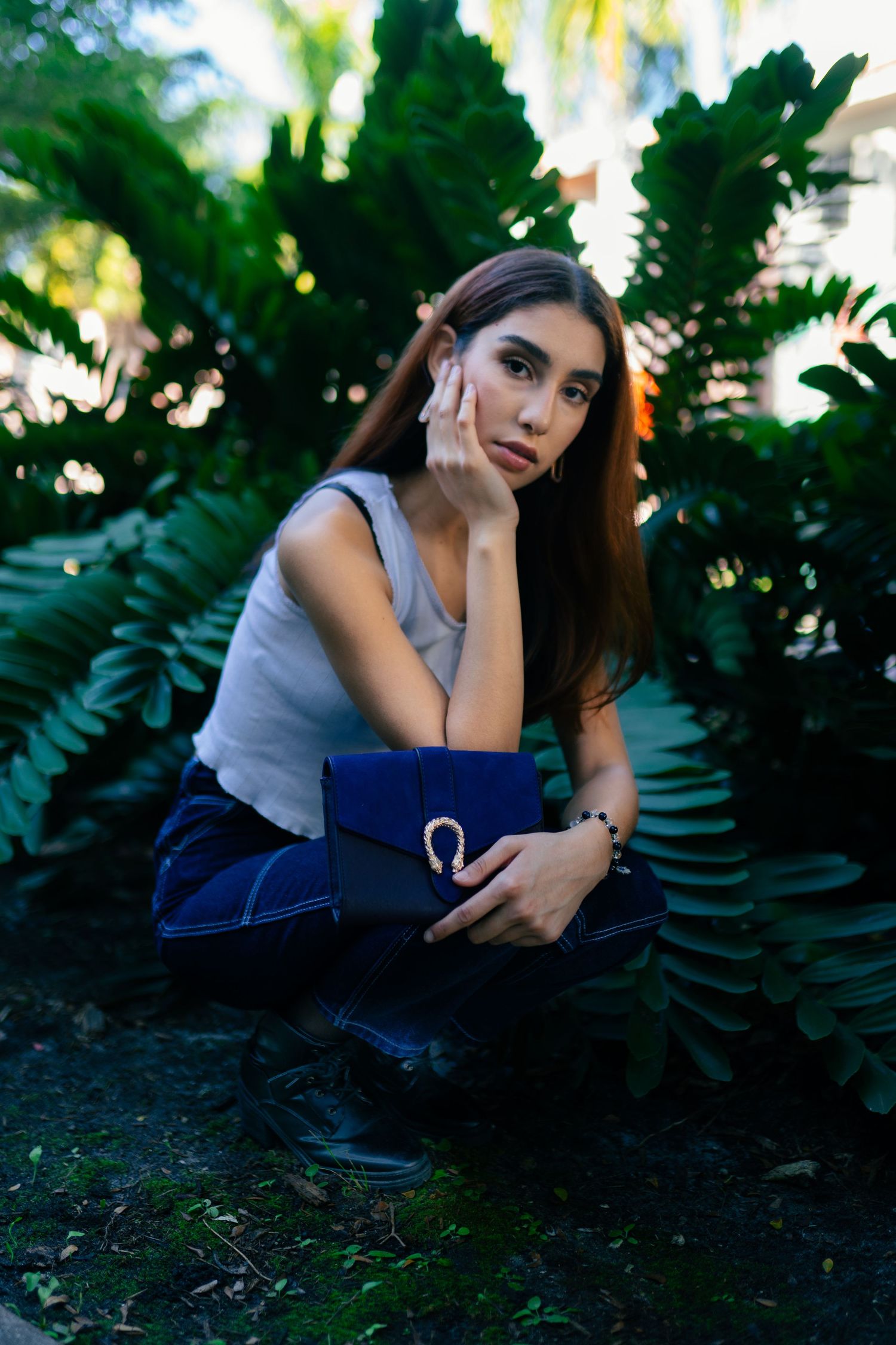 Woman in gray tank top sitting outdoors against lush tropical foliage with a blue book at sunset.