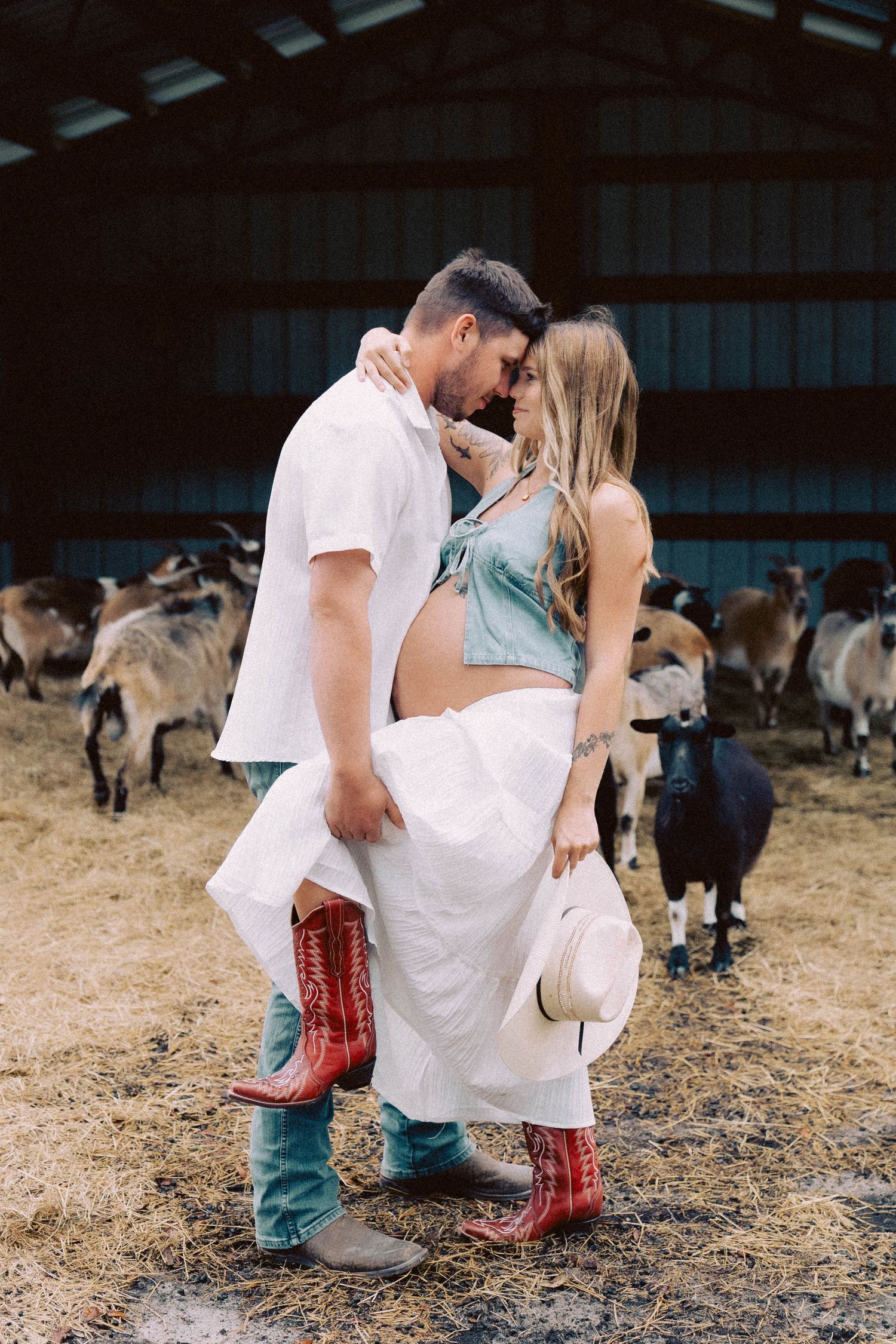 Pregnant couple embracing in a rustic barn surrounded by goats, wearing cowboy boots and casual country attire.