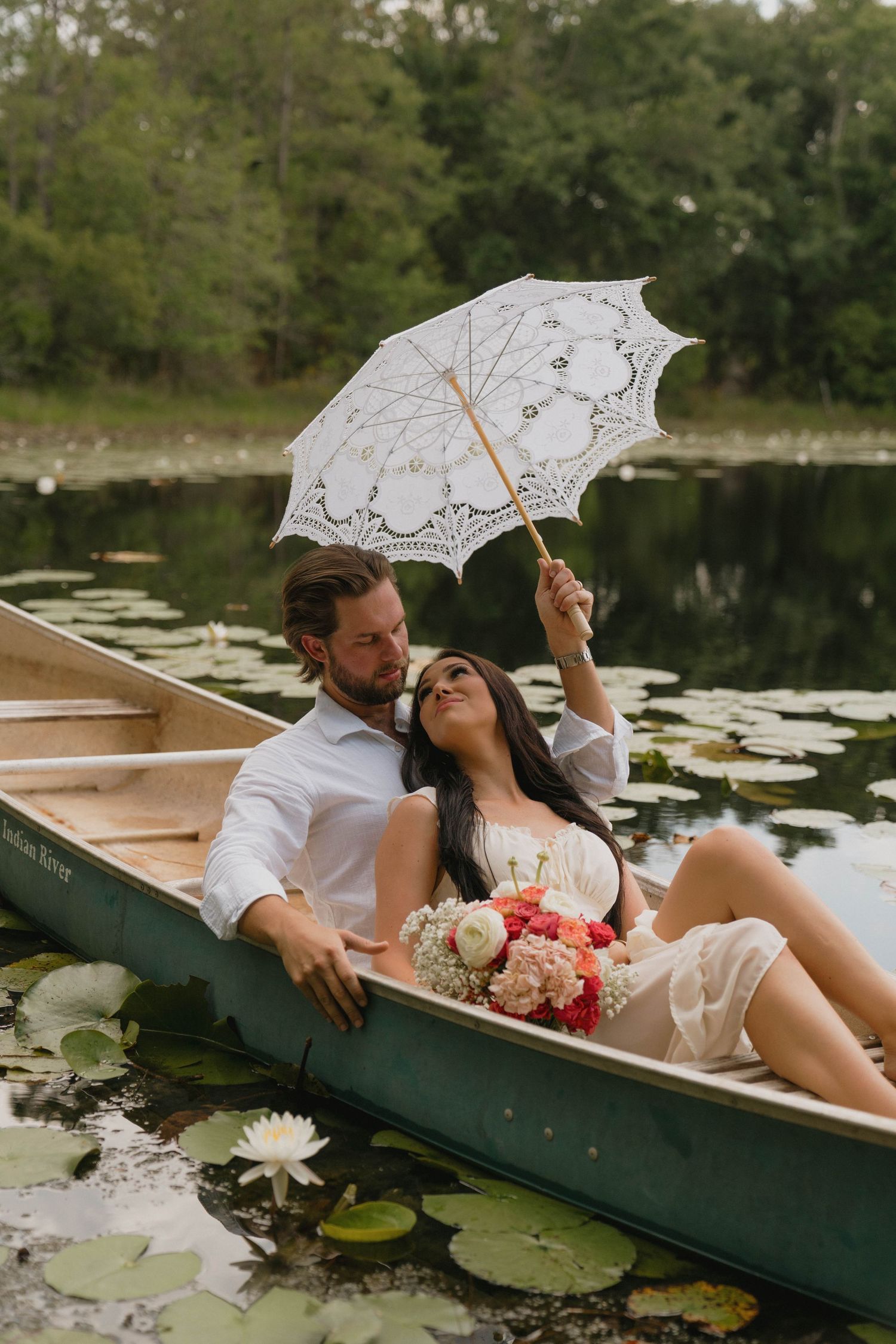 A couple in a canoe on a lily pad lake, bride holding a lace parasol and bouquet, groom embracing her.