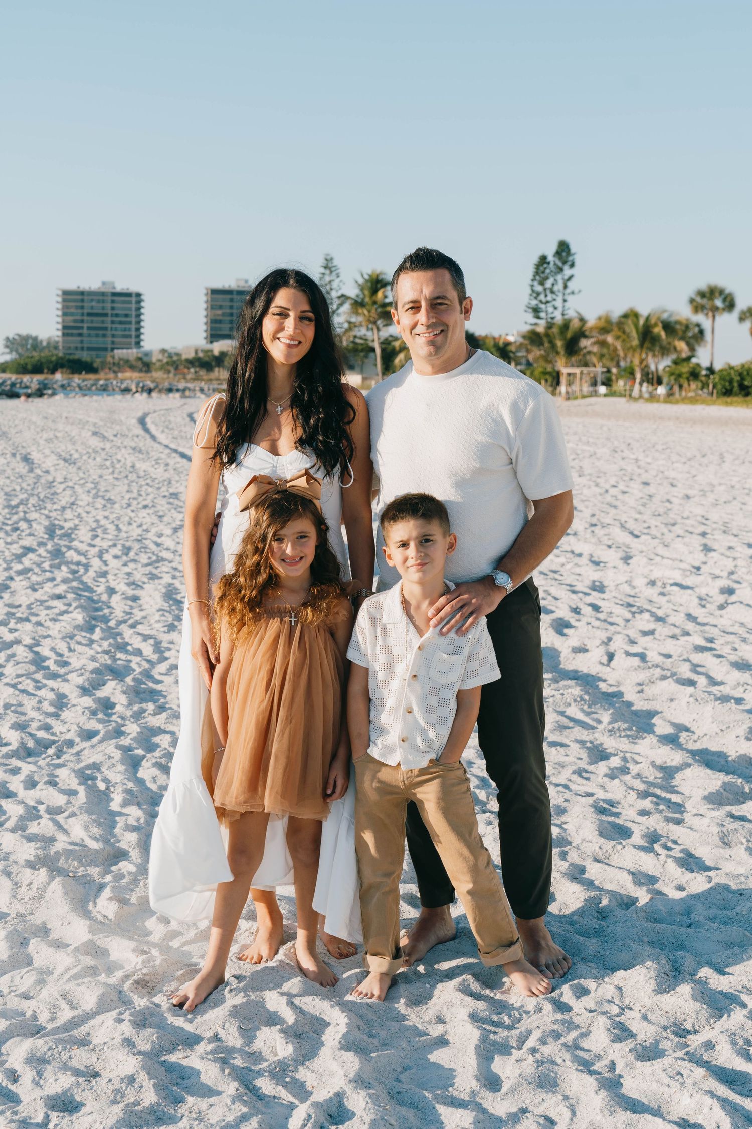 Happy family of four posing together on a white sandy beach with palm trees and buildings in the background.