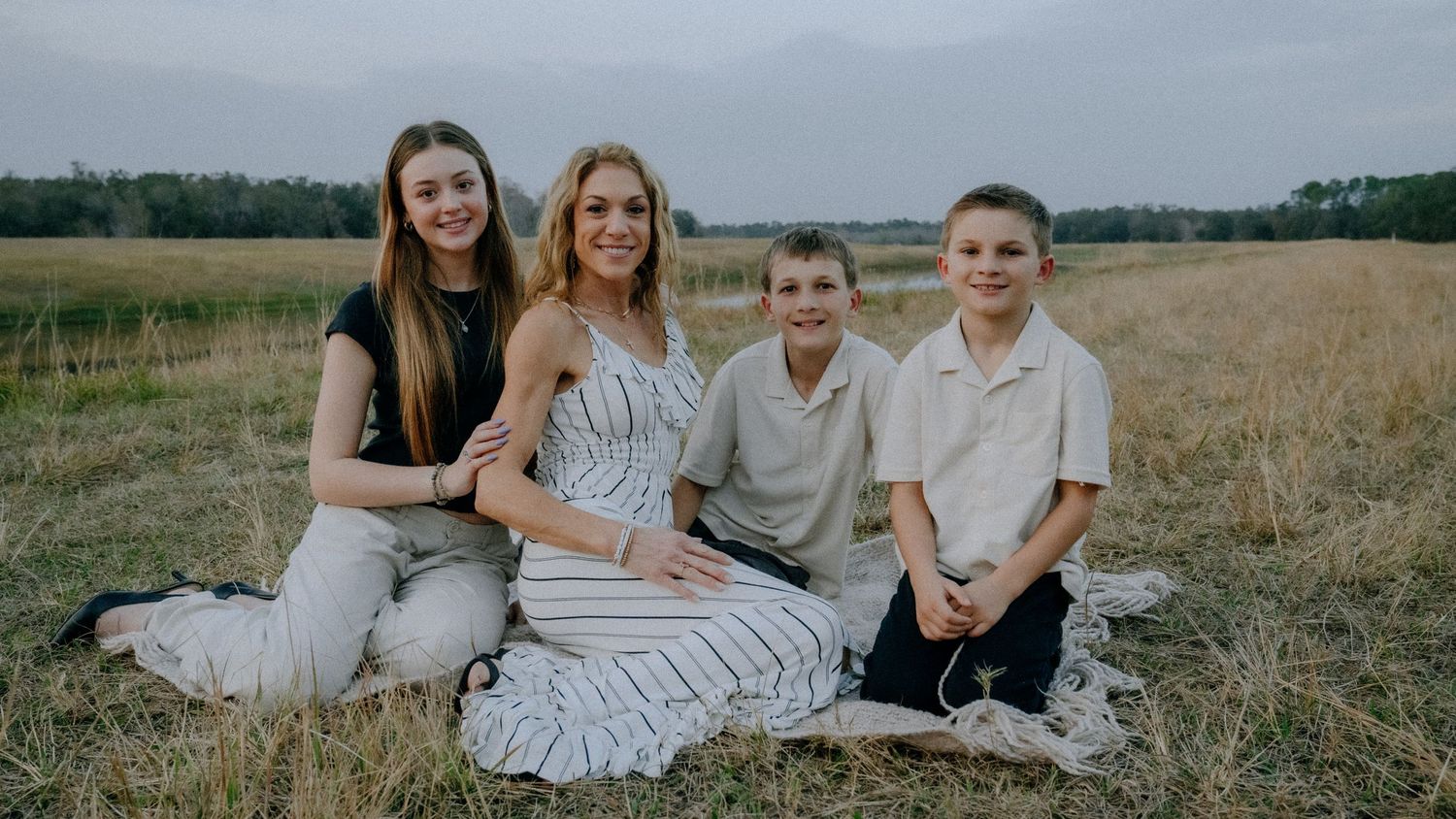 A family group sits together on a blanket in a golden field during sunset with trees in the background.