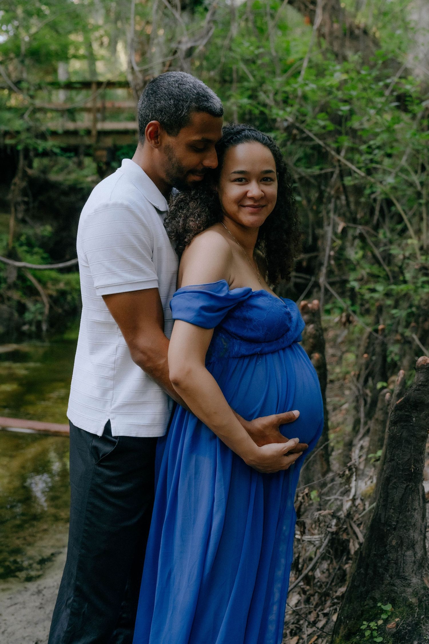 Expectant couple shares tender moment by water's edge with woman wearing flowing blue maternity gown.