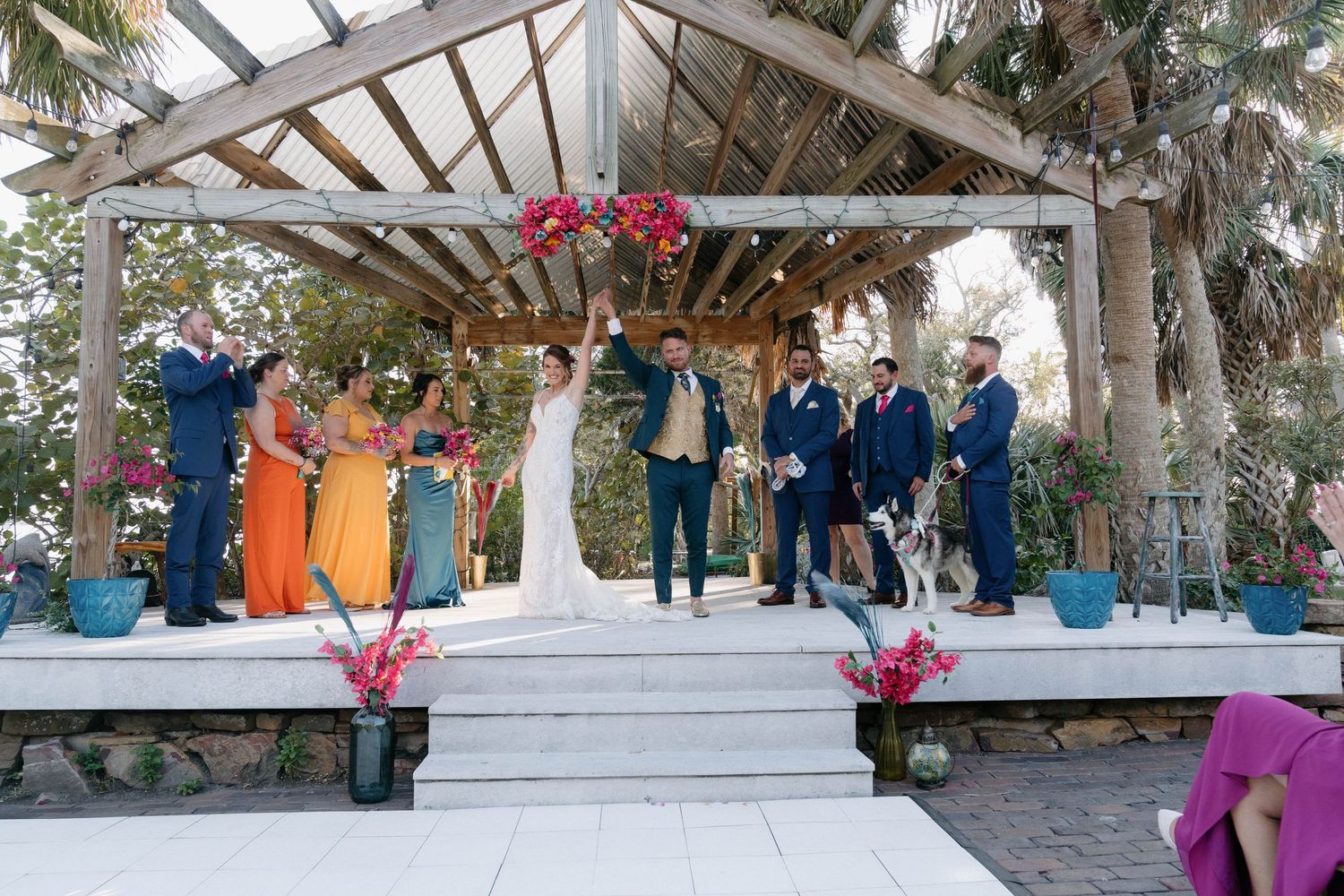 Wedding party celebrating under a rustic pergola with colorful bridesmaid dresses and floral arrangements at an outdoor ceremony.