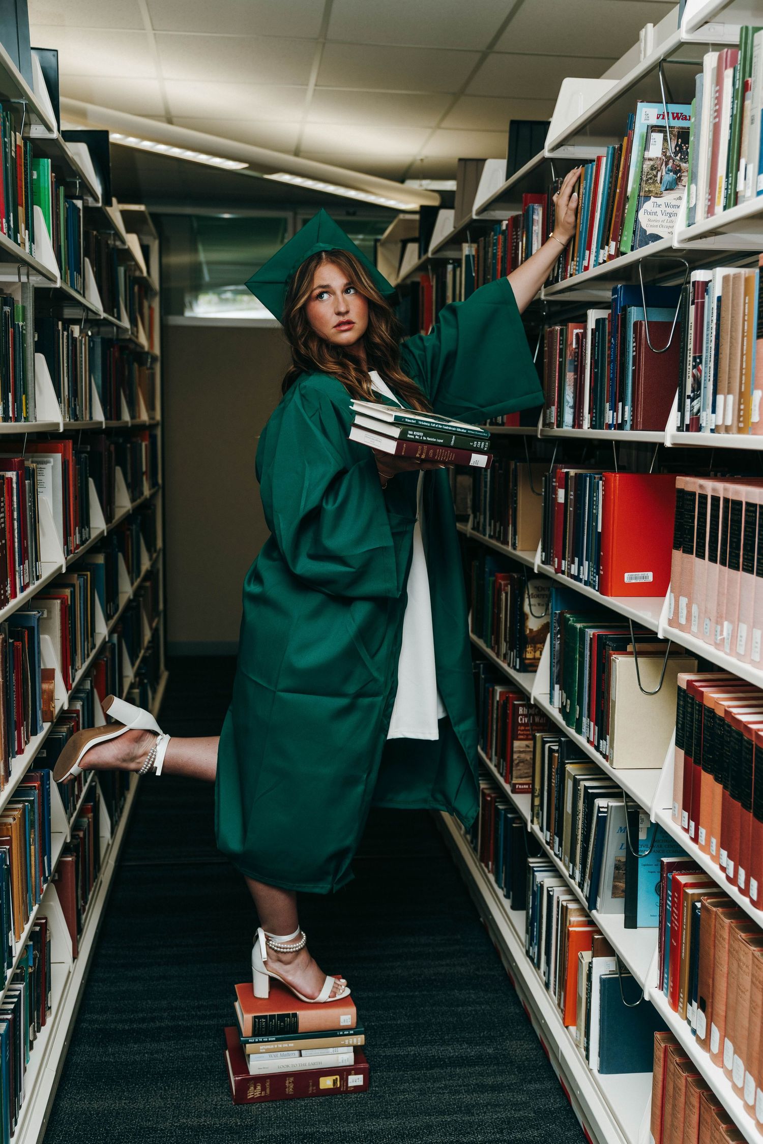 A young woman in a green graduation cap and gown stands on books between library shelves, holding a stack of books.