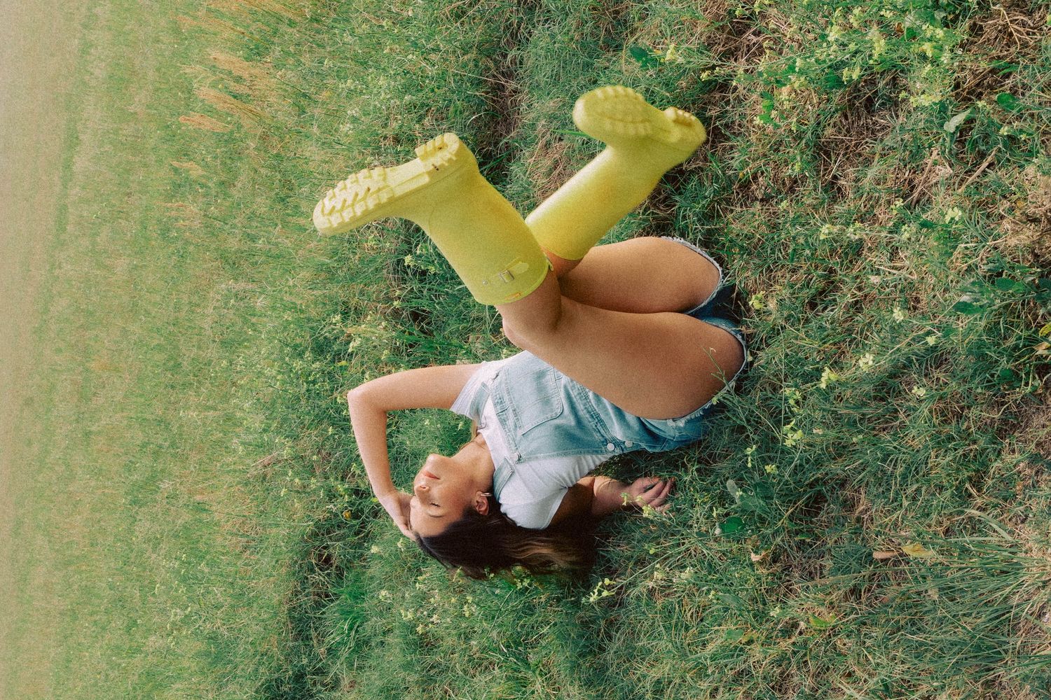 A young woman in yellow rain boots and denim shorts lies playfully on green grass with legs raised in the air.