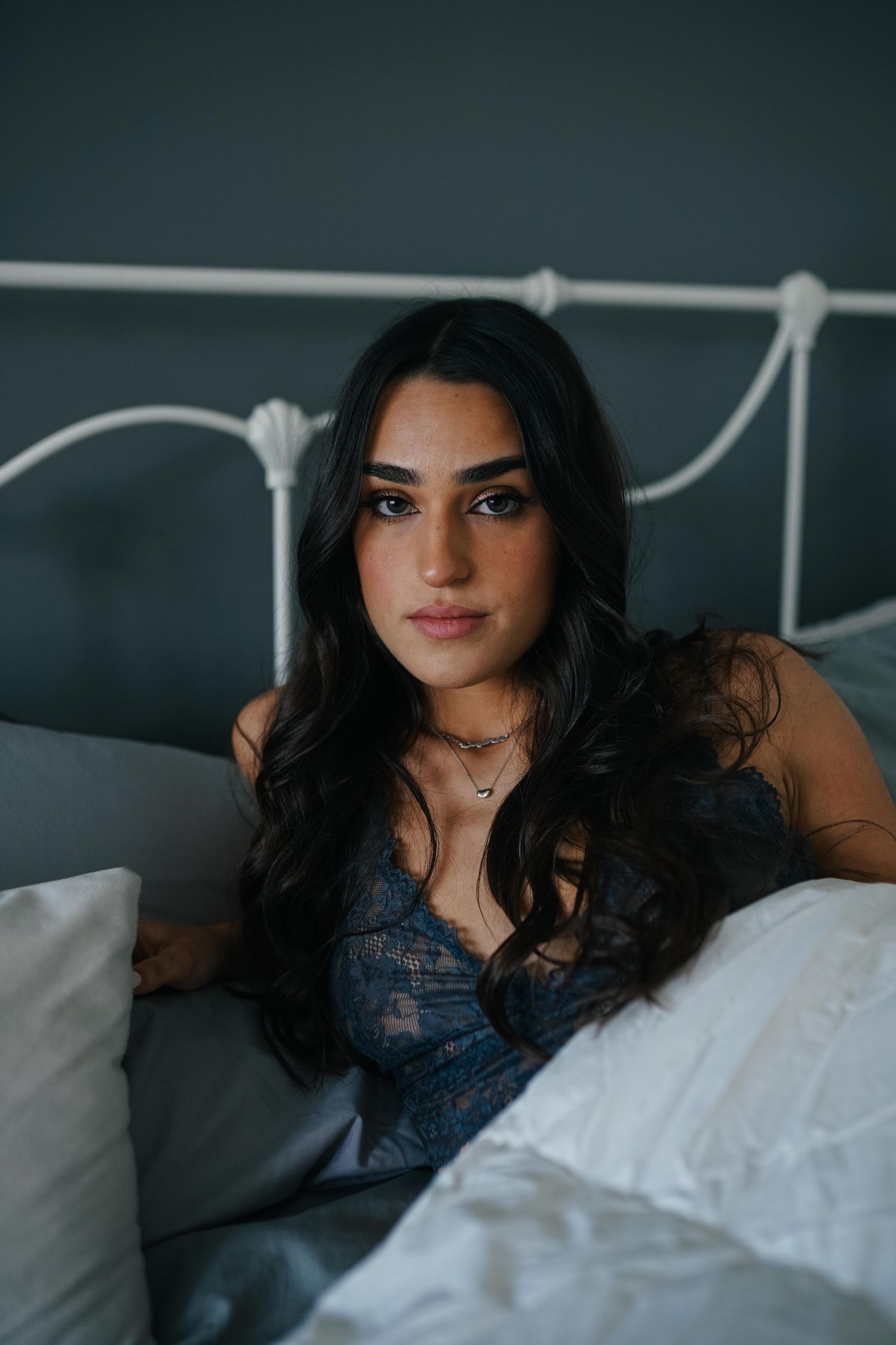 Atmospheric portrait of a woman with long dark hair reclining on a bed with white linens and a metal headboard.