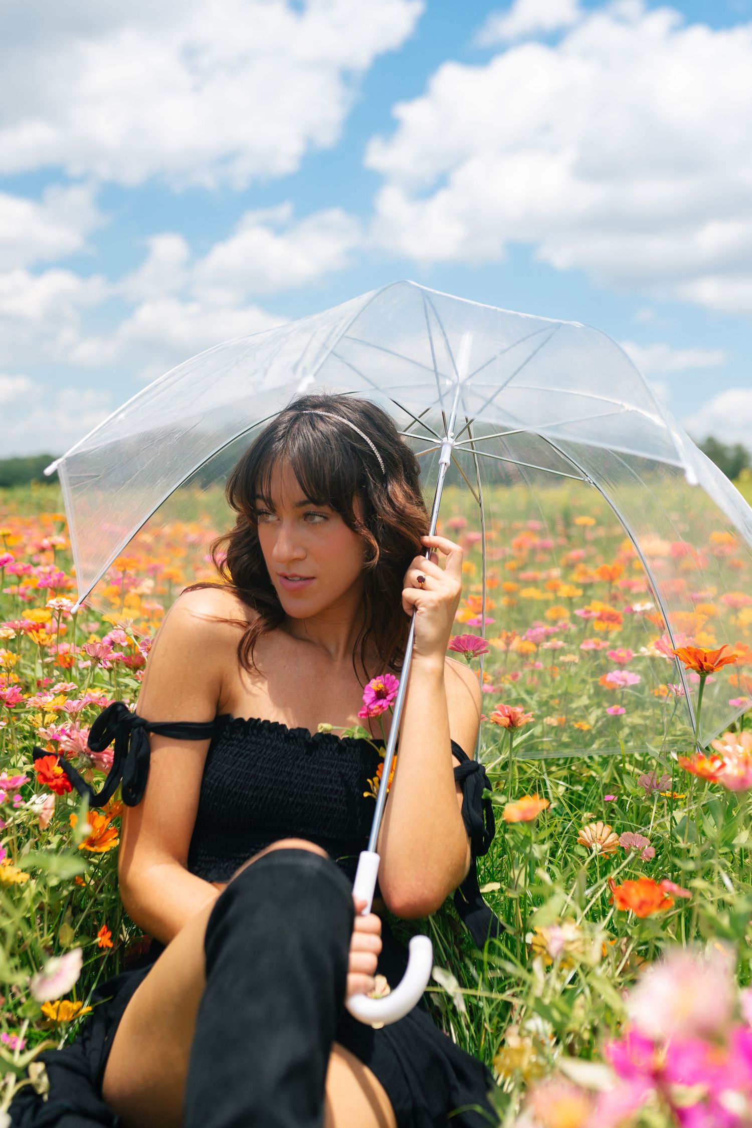 Someone holding a clear umbrella poses in a vibrant field of pink and orange wildflowers under a blue cloudy sky.